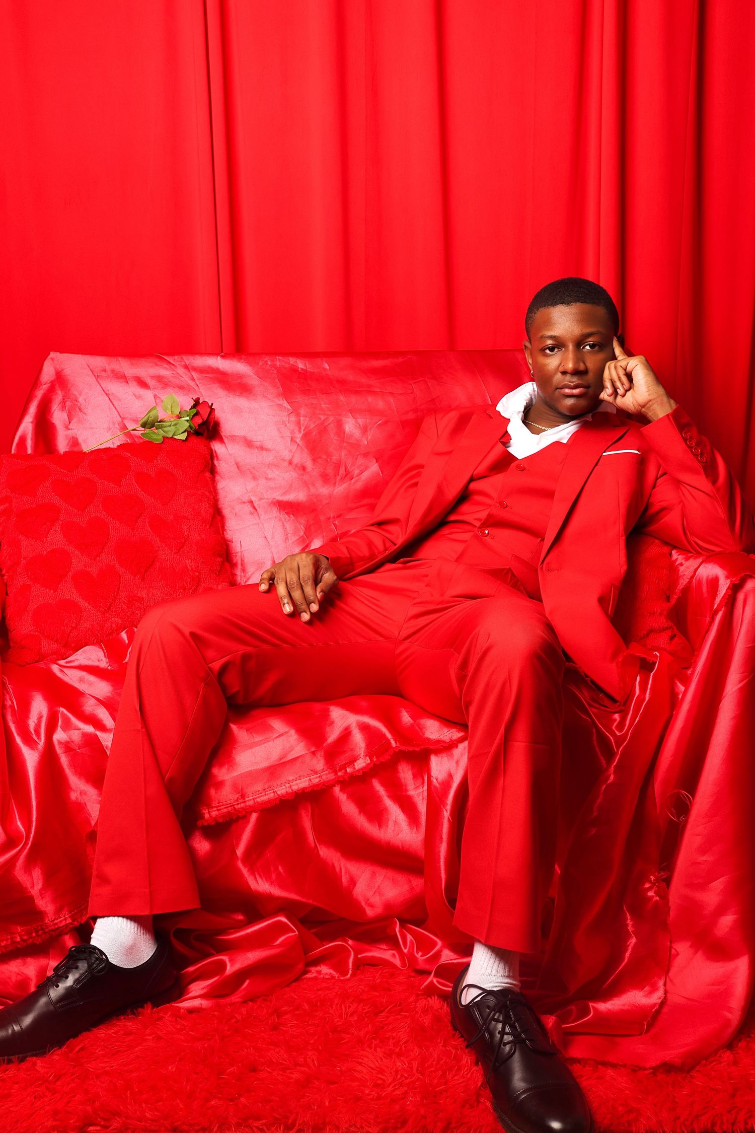 Young man in red suit relaxing on red satin sofa with rose, against red curtain backdrop.