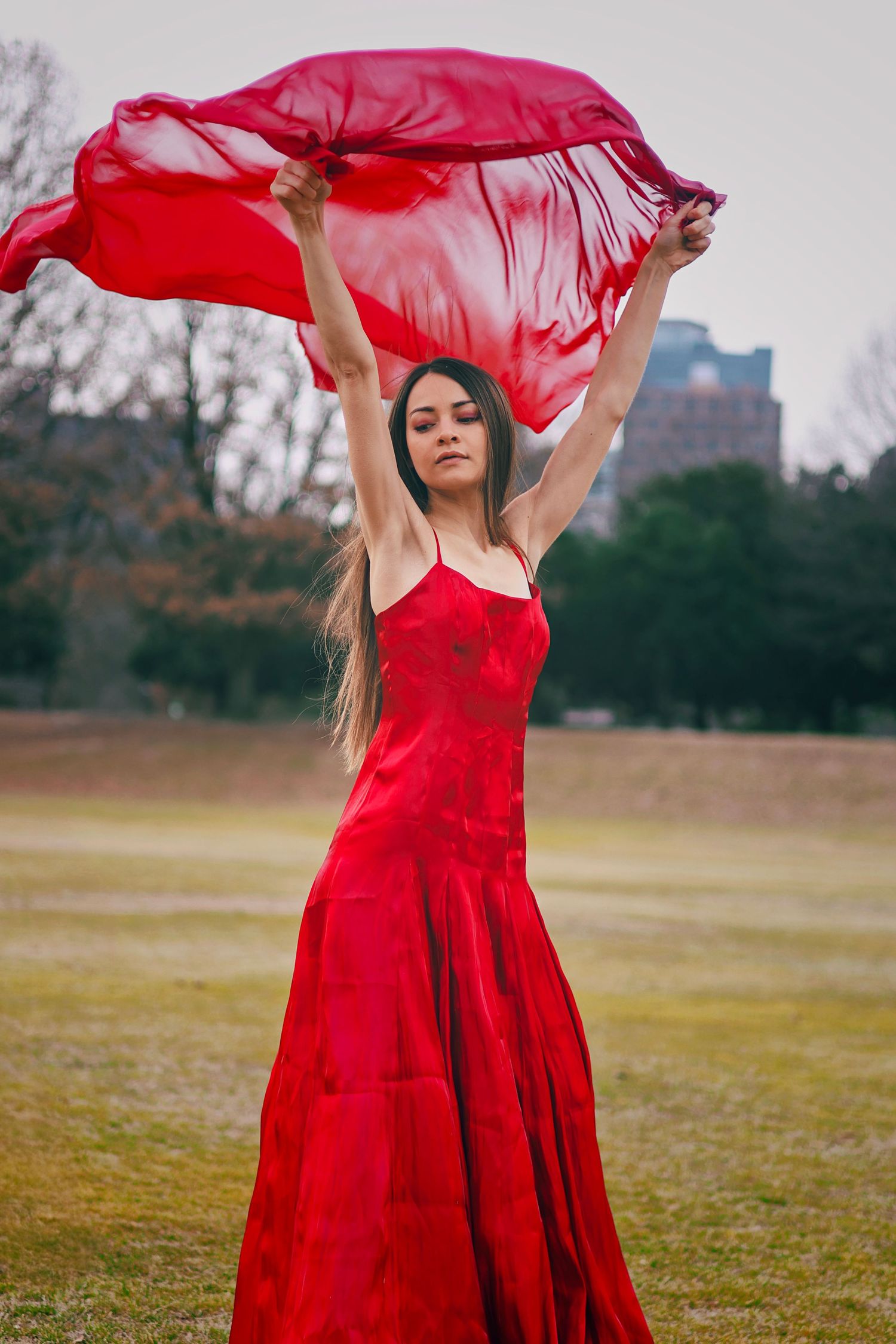 Young woman in red spaghetti strap dress raising red scarf overhead in park.