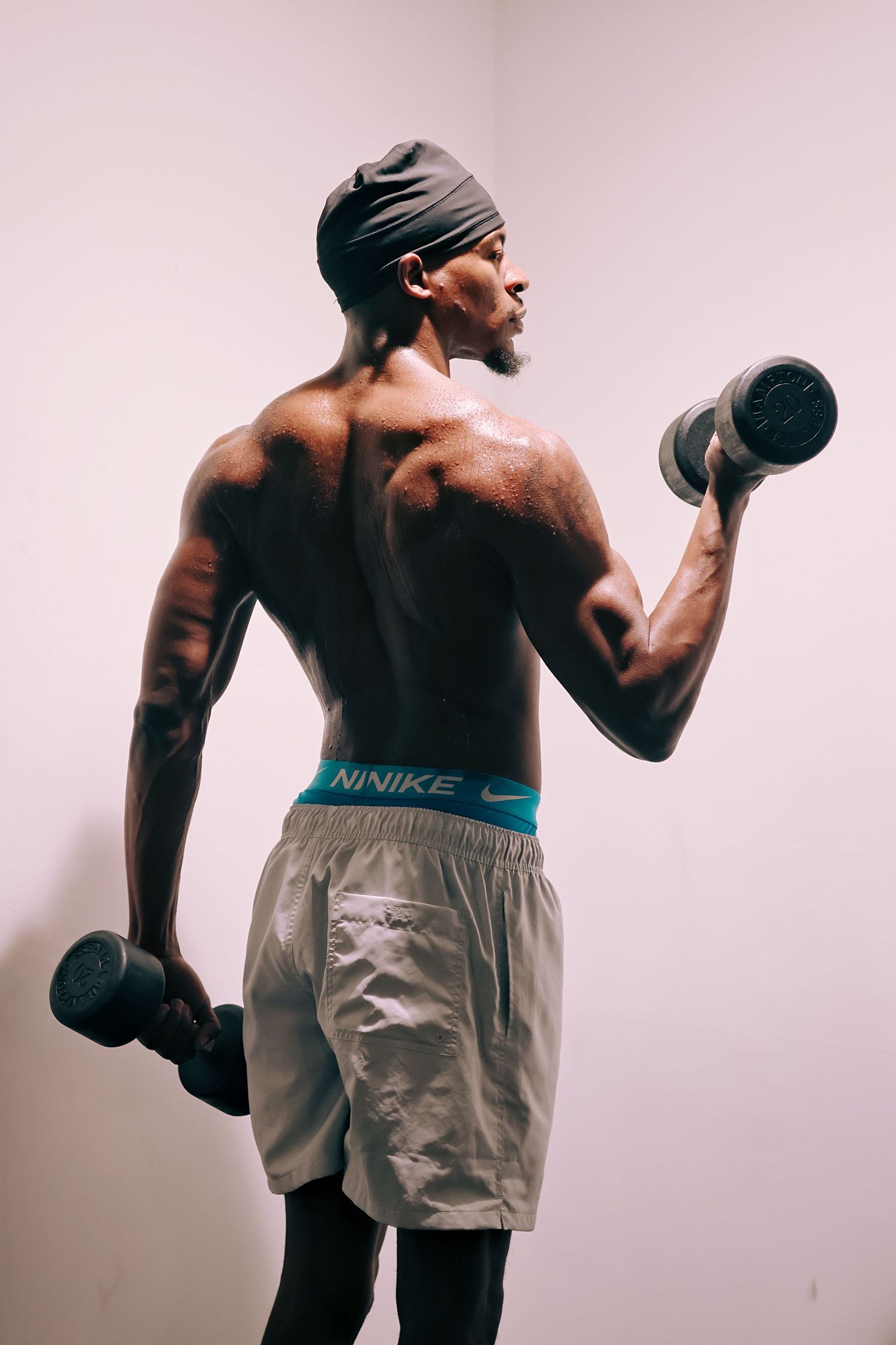 Muscular shirtless man lifting dumbbells wearing Nike underwear and gray shorts, showing defined back muscles during workout.