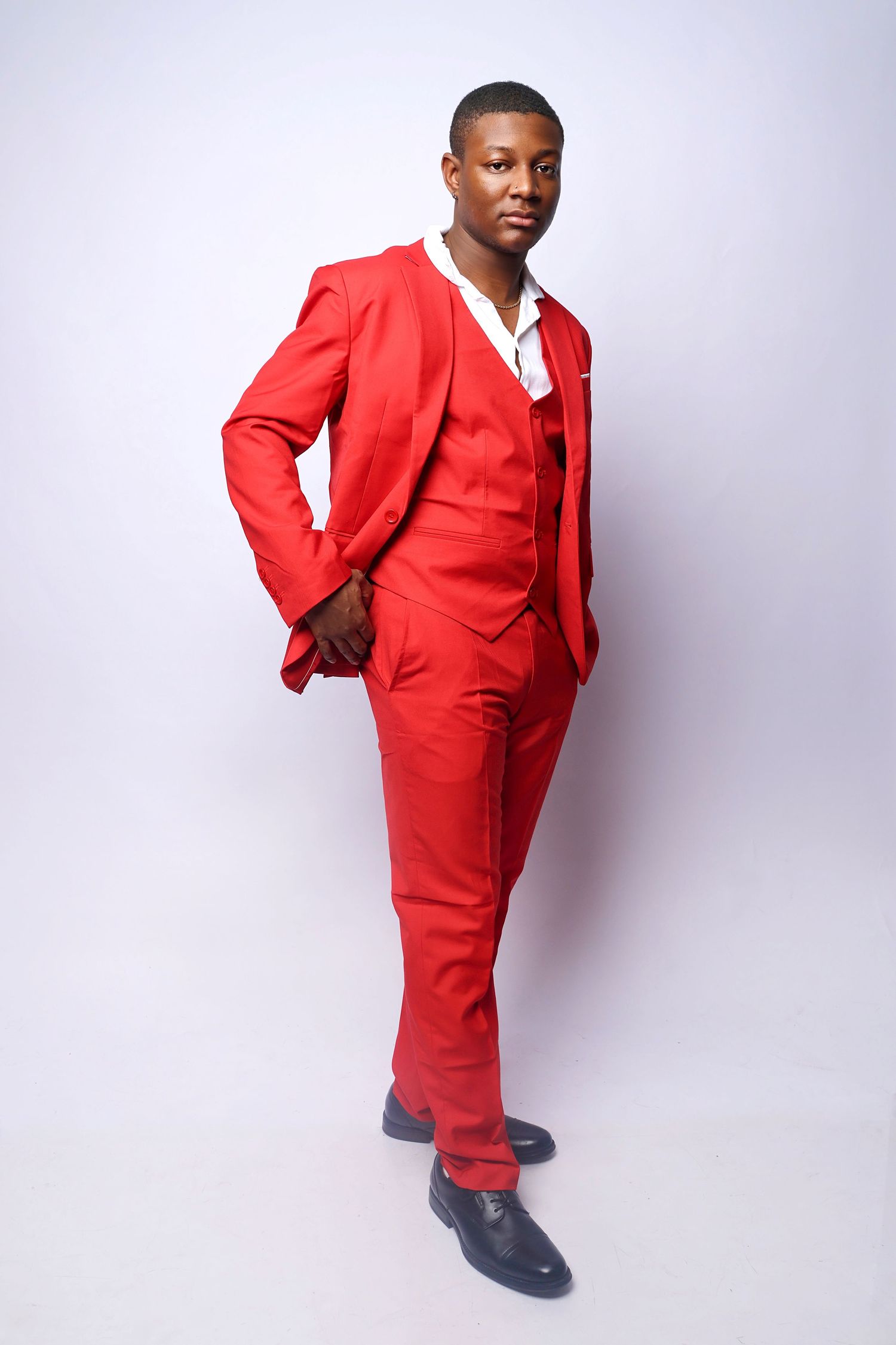 A confident young Black man poses in a bold red three-piece suit with black dress shoes on a white background.