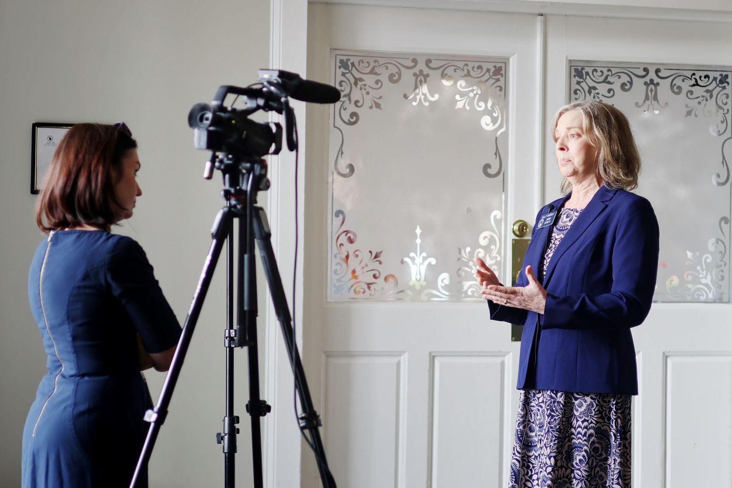 Two women filming a professional video interview indoors, one operating a camera on tripod, other holding notes.