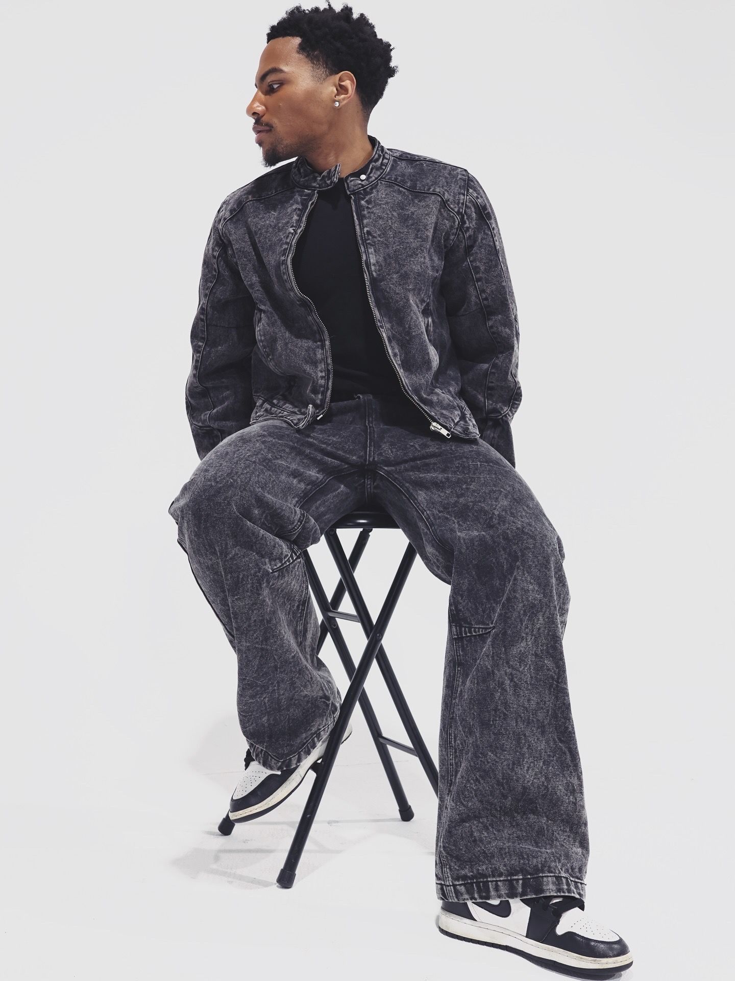 Young Black man seated on a stool wearing acid wash dark denim jacket and wide-leg jeans with Nike sneakers.