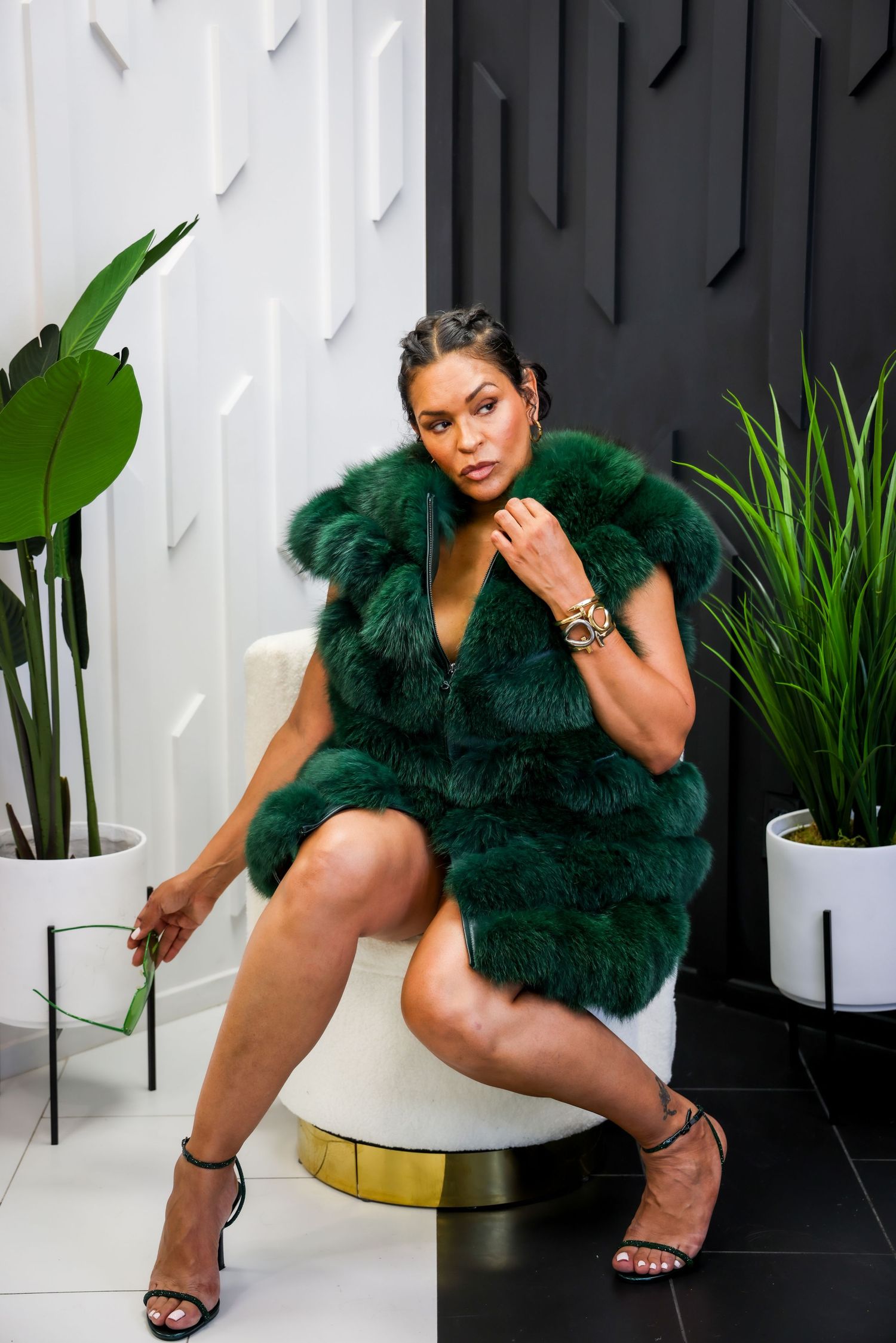 Stylish woman in emerald green faux fur vest seated in white chair, luxury fashion portrait.