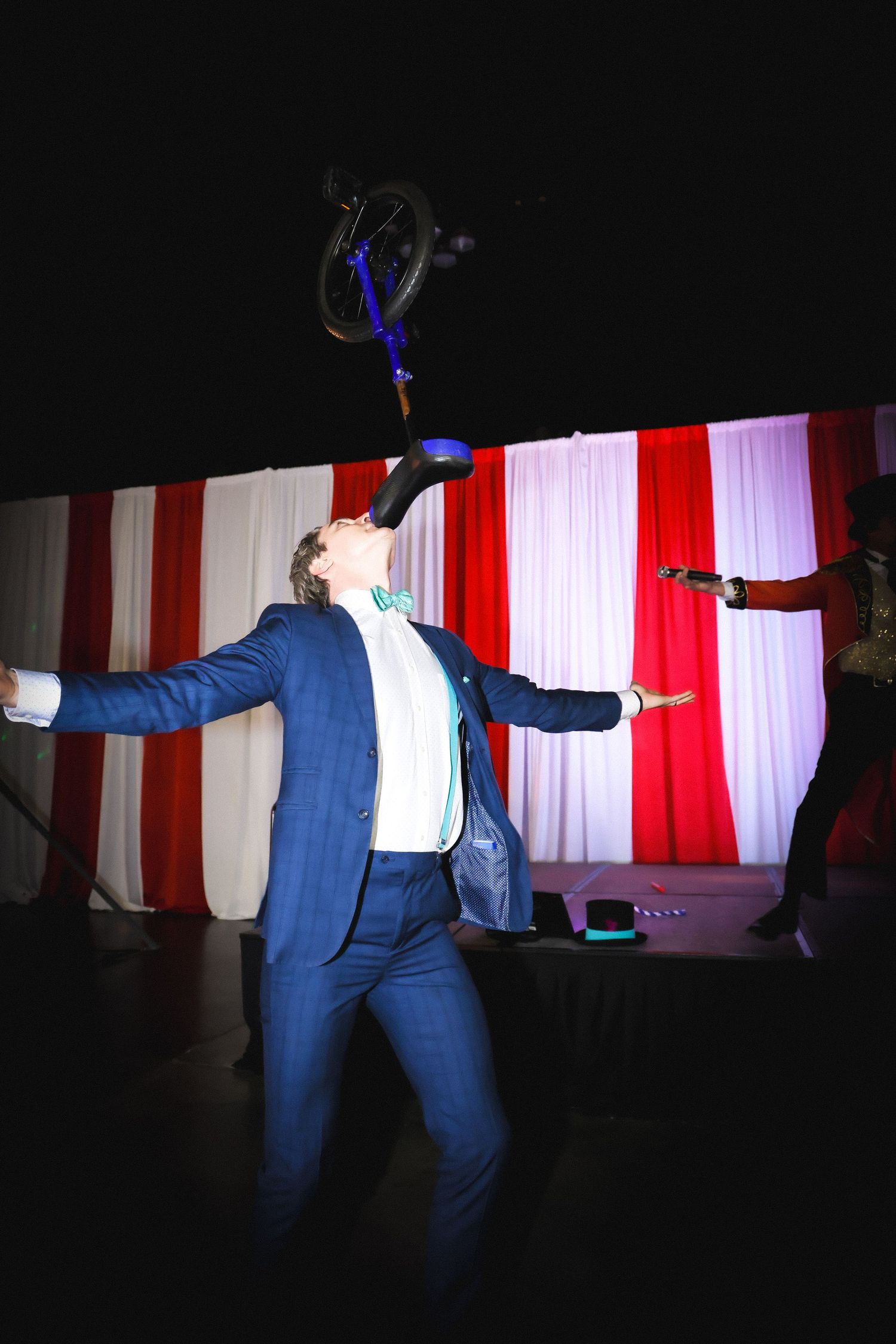 Man in blue suit balancing a unicycle on his chin on stage with red and white curtains backdrop.