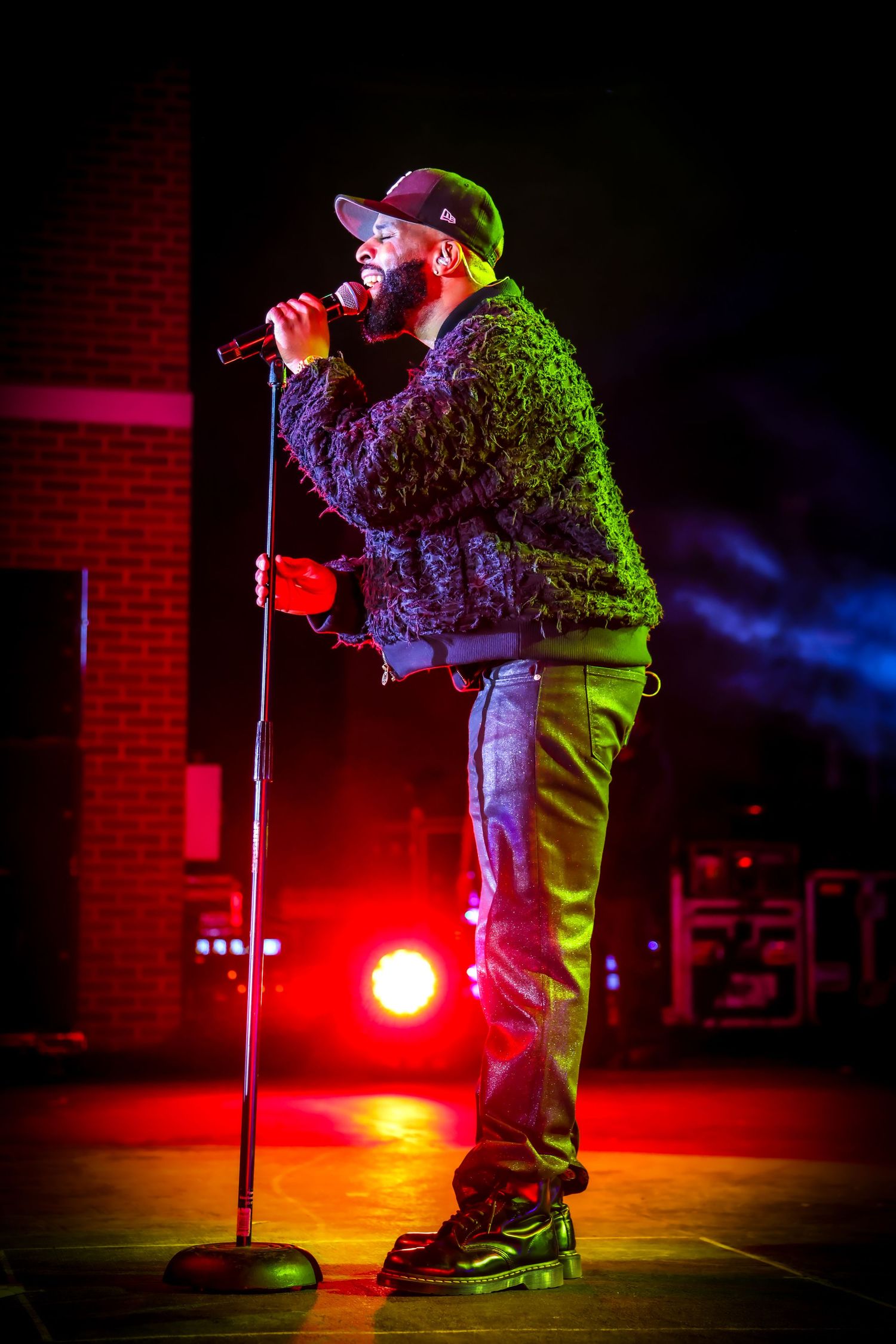 A male singer performs on stage holding a microphone on a stand, wearing a fuzzy jacket and cap under dramatic red stage lighting.