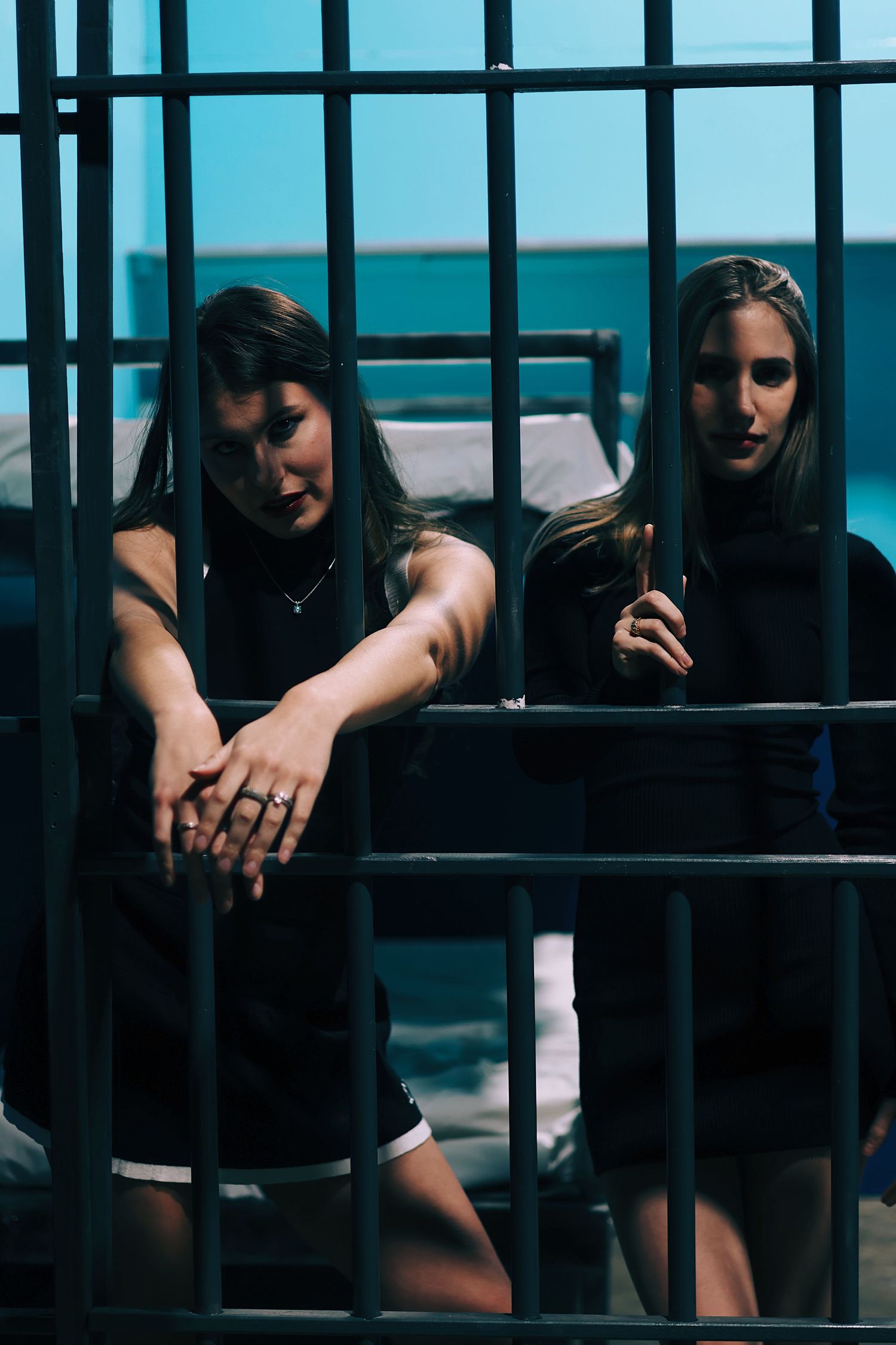 Two women in black dresses behind jail cell bars in dramatic blue-lit prison scene.