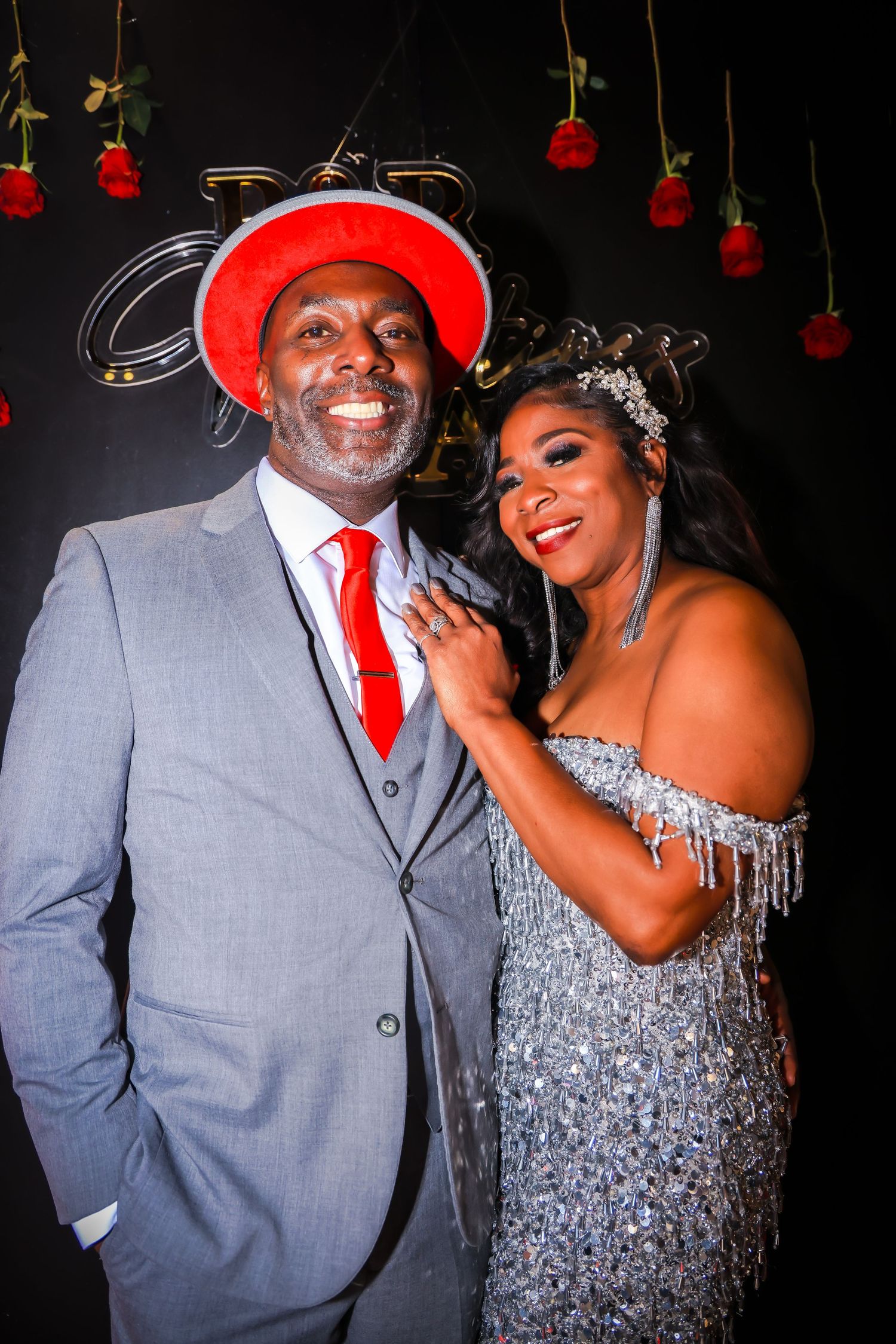 Elegant couple posing at formal event, man in gray suit with red hat and tie, woman in silver beaded gown with crown.