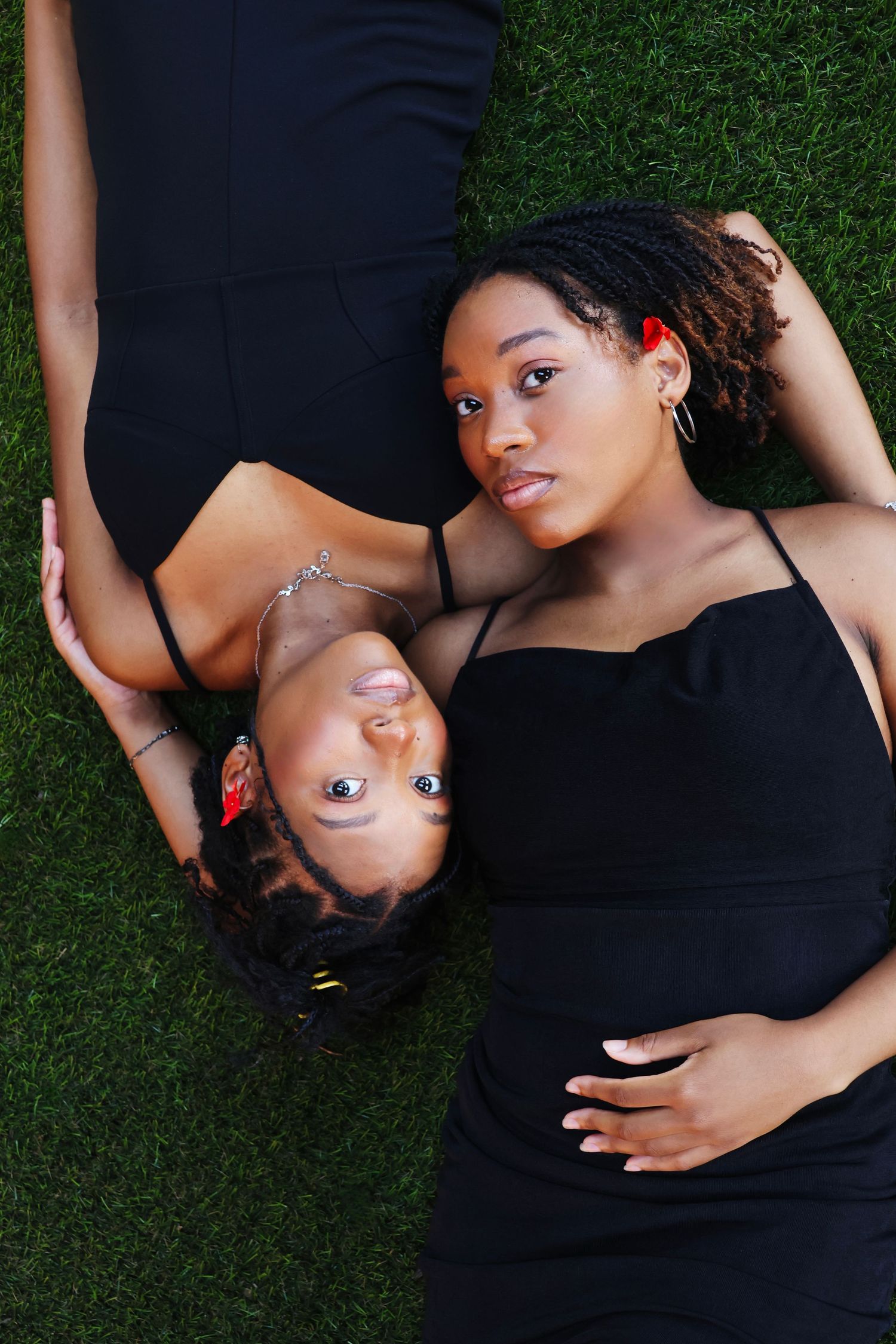 Two young Black women in black dresses lying on green grass, looking up at the camera with relaxed expressions.