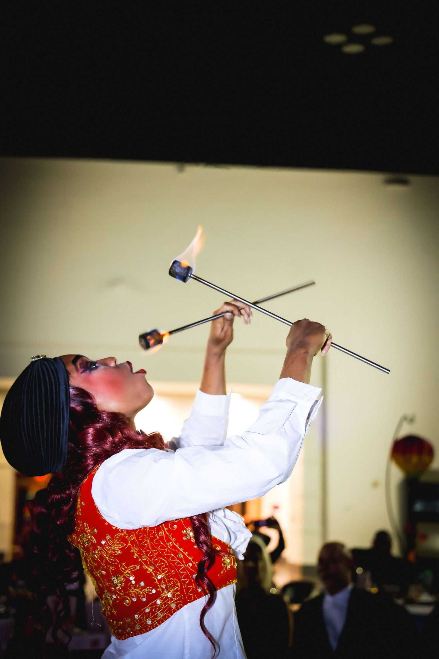 A fire performer in traditional red and white costume balances a flaming baton during an indoor cultural show.