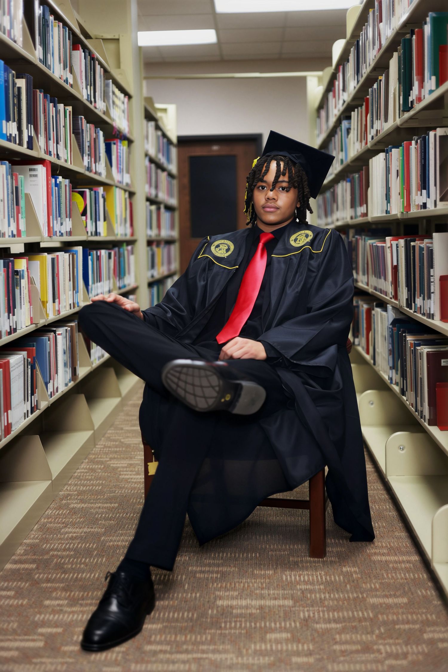 Graduate in black cap and gown with red tie sits confidently between library bookshelves for graduation portrait.