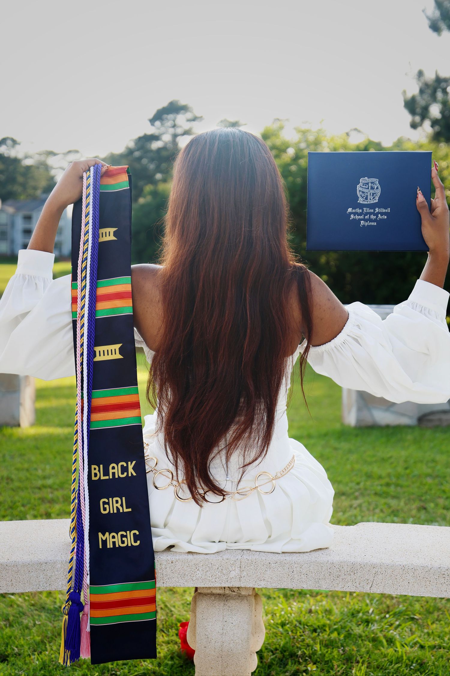 Graduate holding diploma and Black Girl Magic kente stole, seated outdoors in white dress with long brown hair.