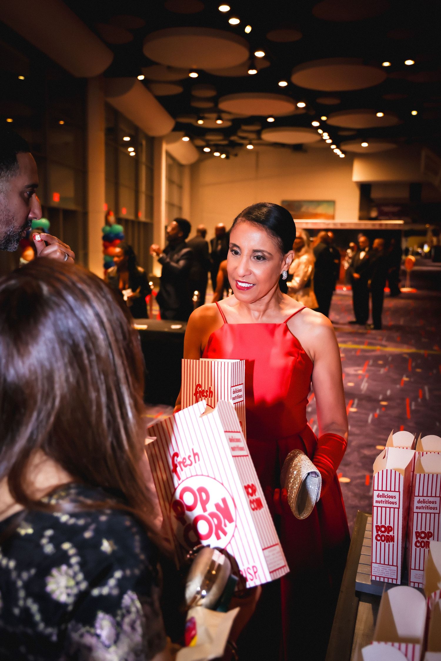 Woman in red gown smiling while holding popcorn bags at an elegant event.