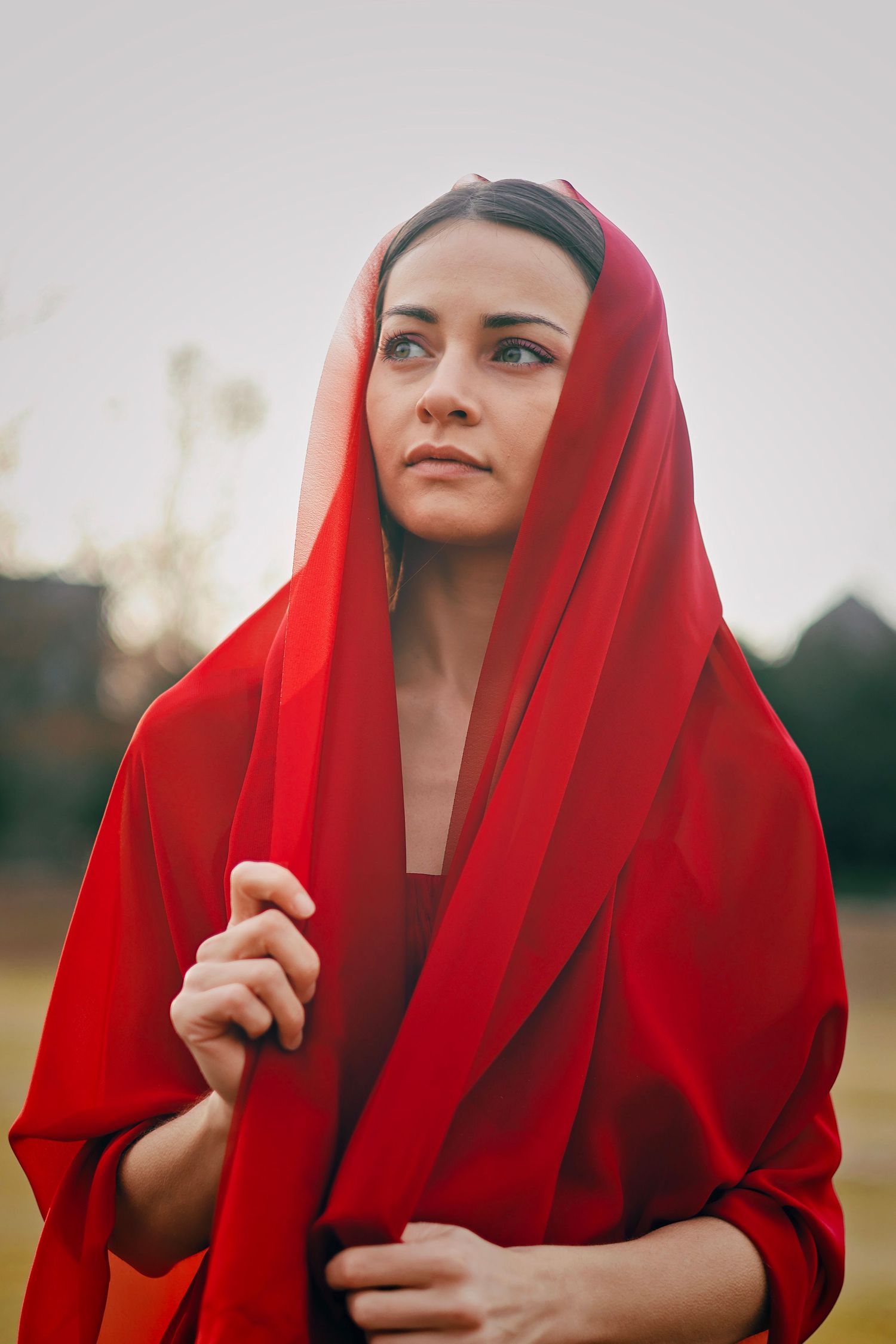 Woman in red headscarf and wrap gazes upward outdoors in a serene, contemplative pose.