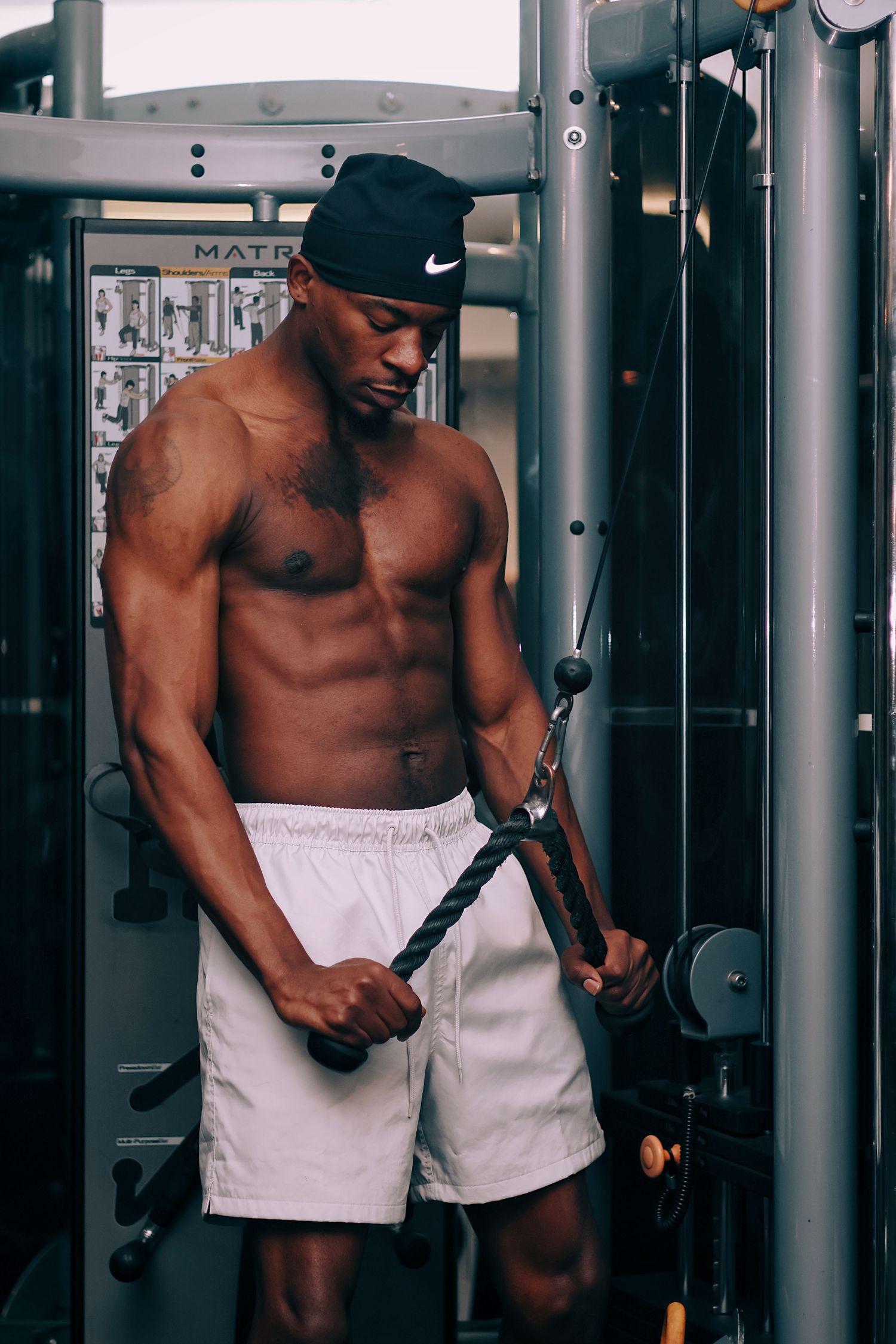 Muscular shirtless Black man in white shorts and Nike cap doing cable tricep pushdowns at a Matrix gym machine.