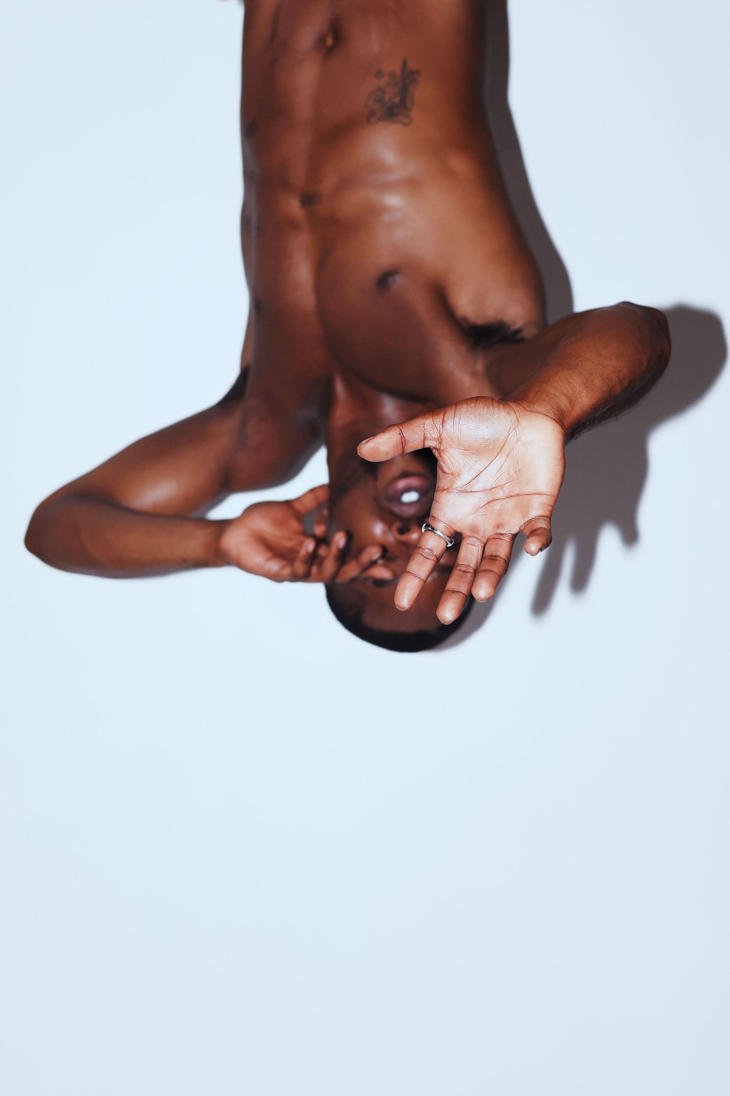 Shirtless Black man lying back with tattooed torso and outstretched hand on white background.