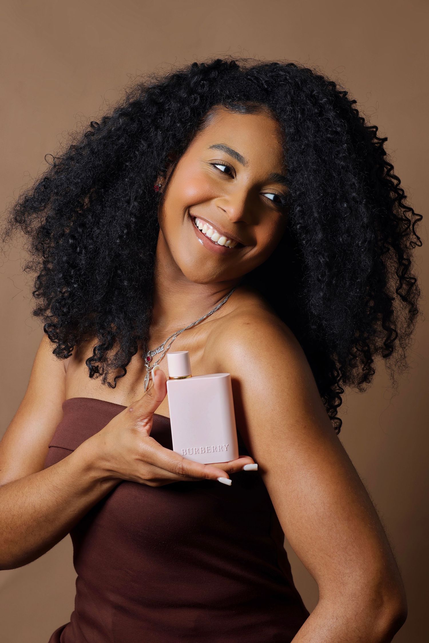 Smiling woman with curly hair holding a pink Burberry perfume bottle, wearing a brown strapless top on beige background.