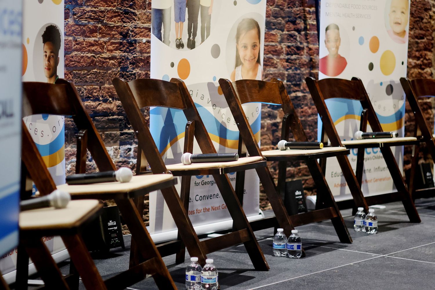 Rows of empty wooden folding chairs arranged in front of banners with portraits at an indoor event venue.