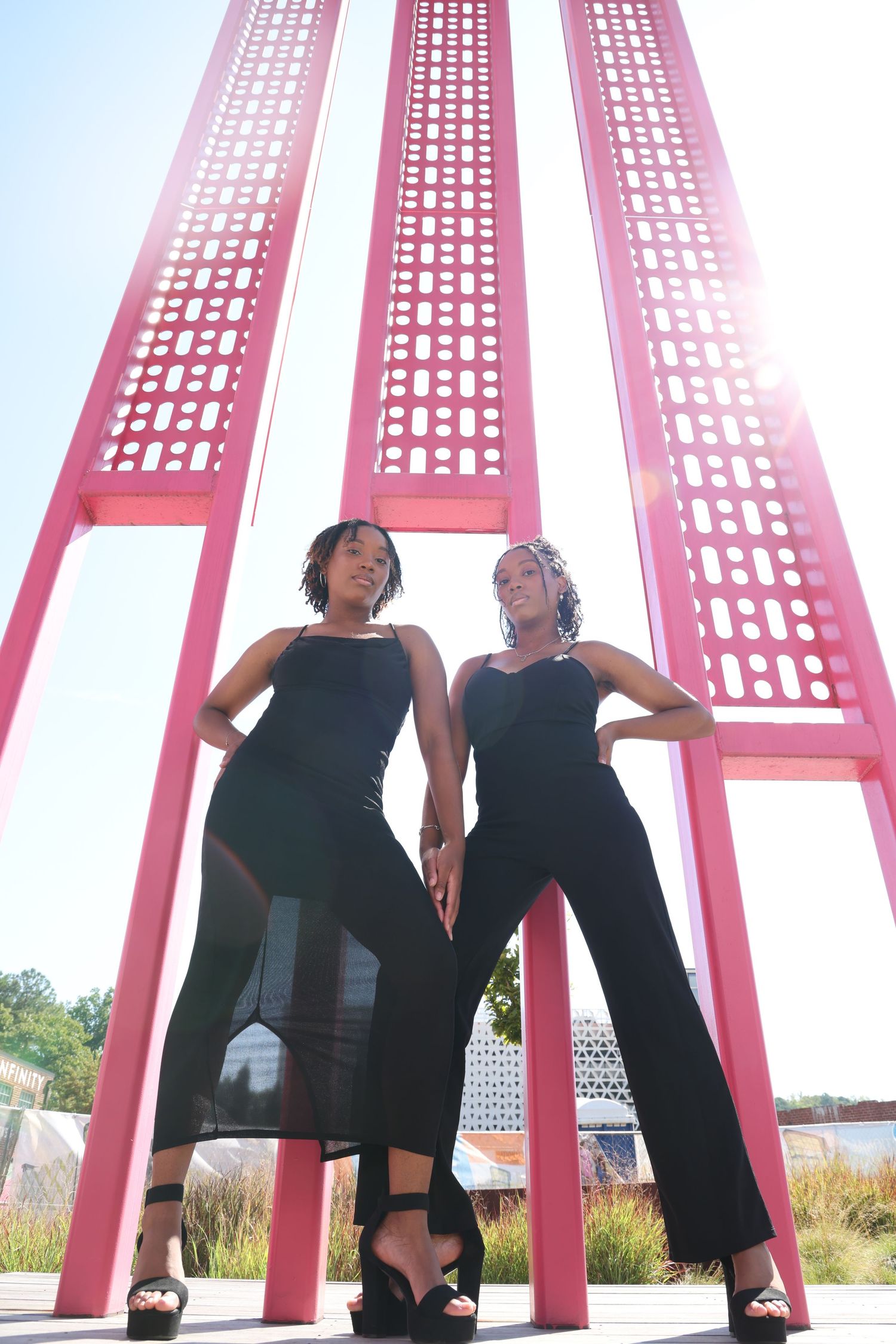 Two women in black outfits pose confidently in front of vibrant pink architectural columns outdoors.