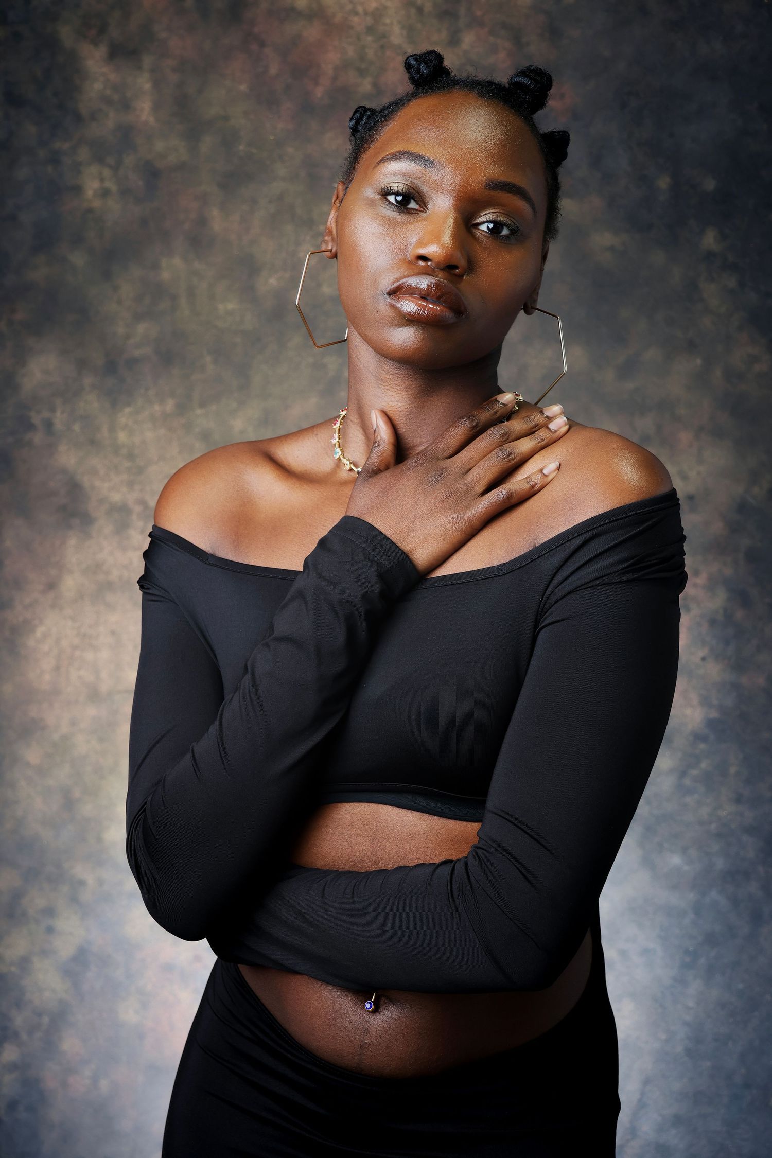 Black woman in off-shoulder black top with hexagonal earrings posing against textured studio backdrop.