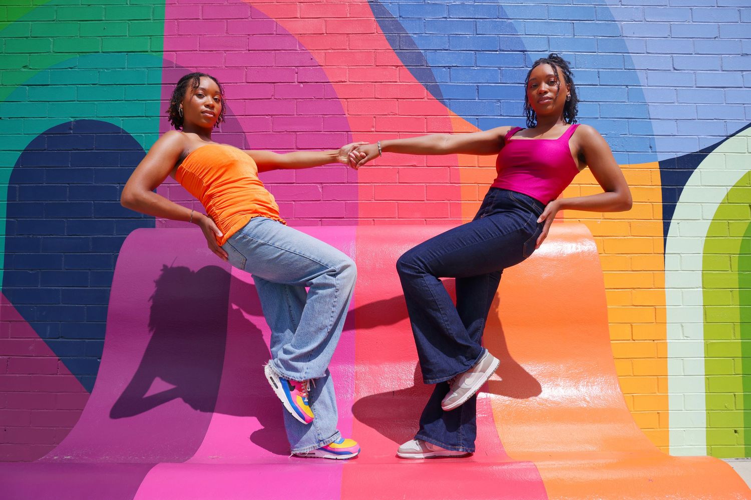 Two women in colorful tops holding hands, posing against vibrant rainbow mural wall.