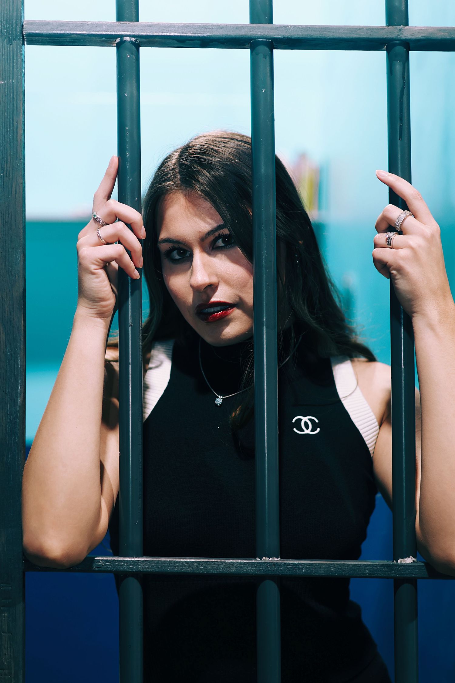 Young woman in black Chanel top gripping metal jail bars, looking directly at camera with intense expression.
