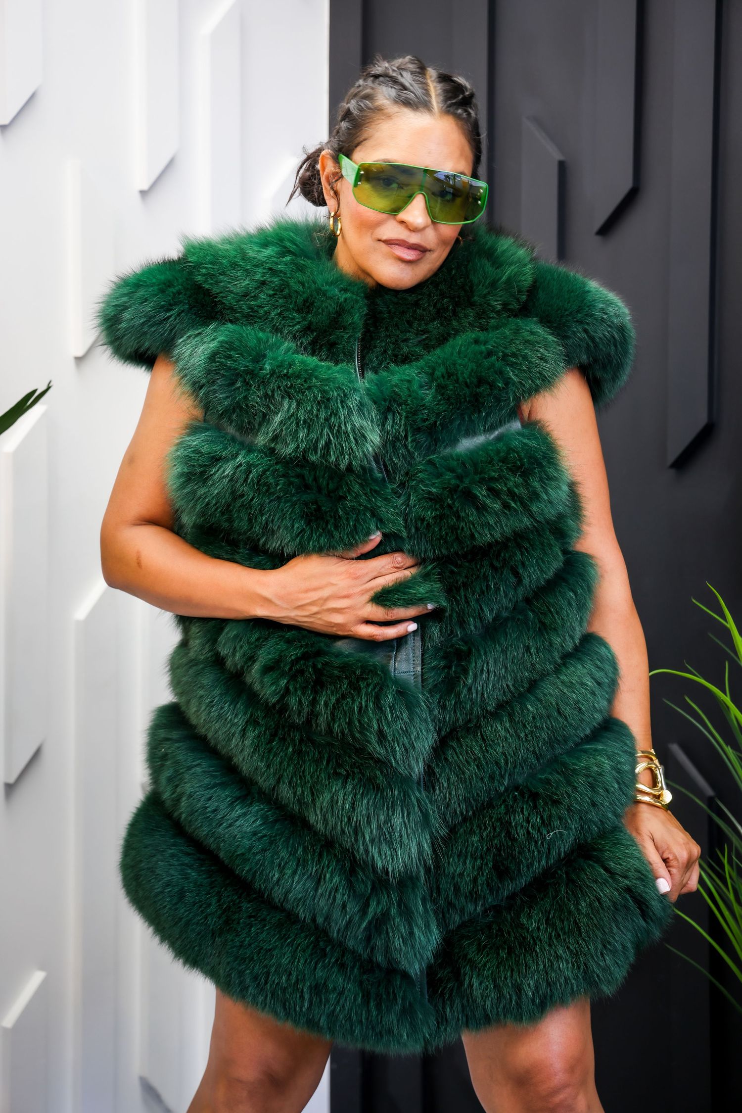 Stylish woman wearing oversized emerald green fur vest with matching sunglasses standing at doorway.