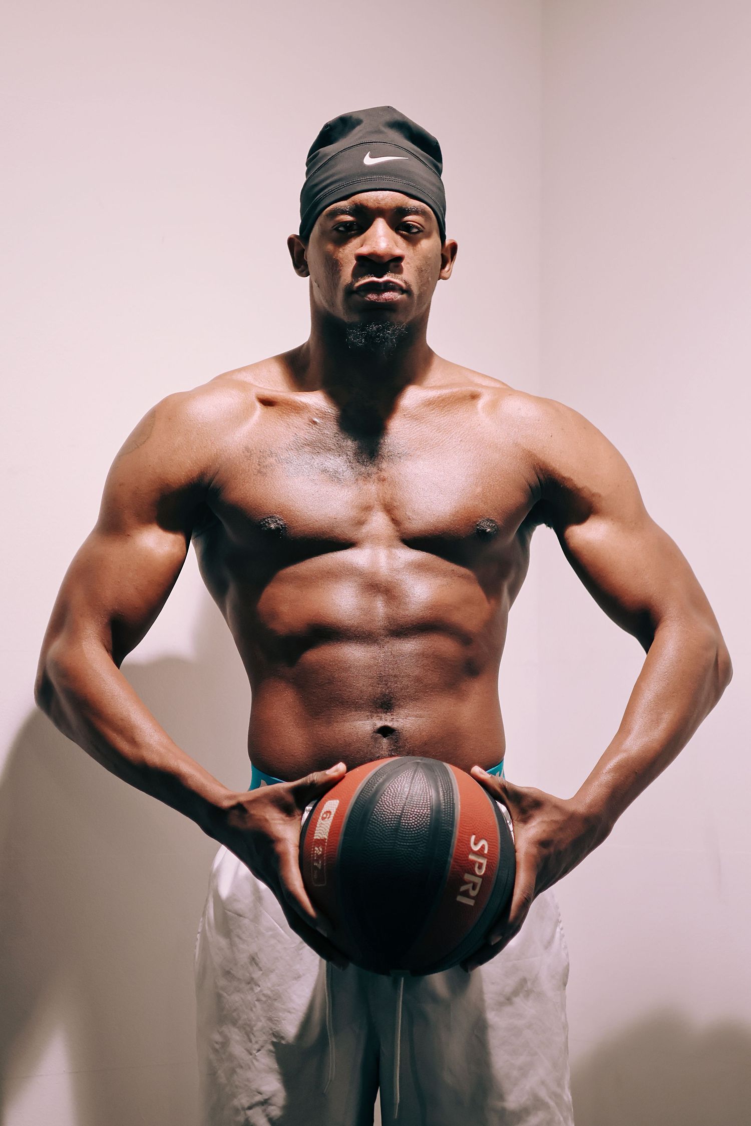 Muscular shirtless Black man holding basketball wearing Nike headband and white shorts.