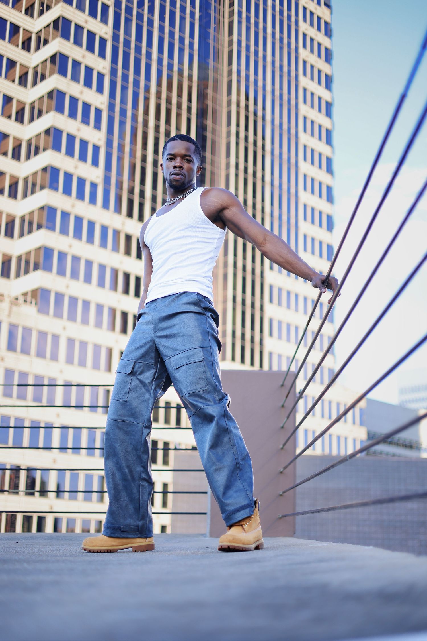 Young Black man in white tank top, cargo jeans, and boots posing confidently in urban cityscape with skyscrapers.