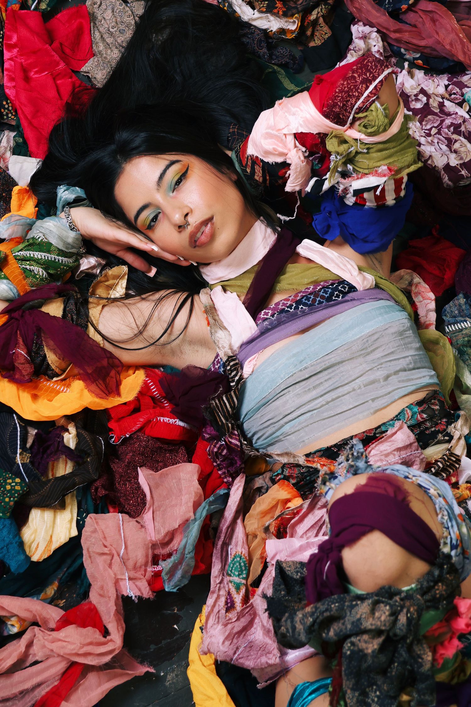 A young woman with dark hair lies among a colorful pile of mixed fabrics and clothing scraps in a fashion editorial.