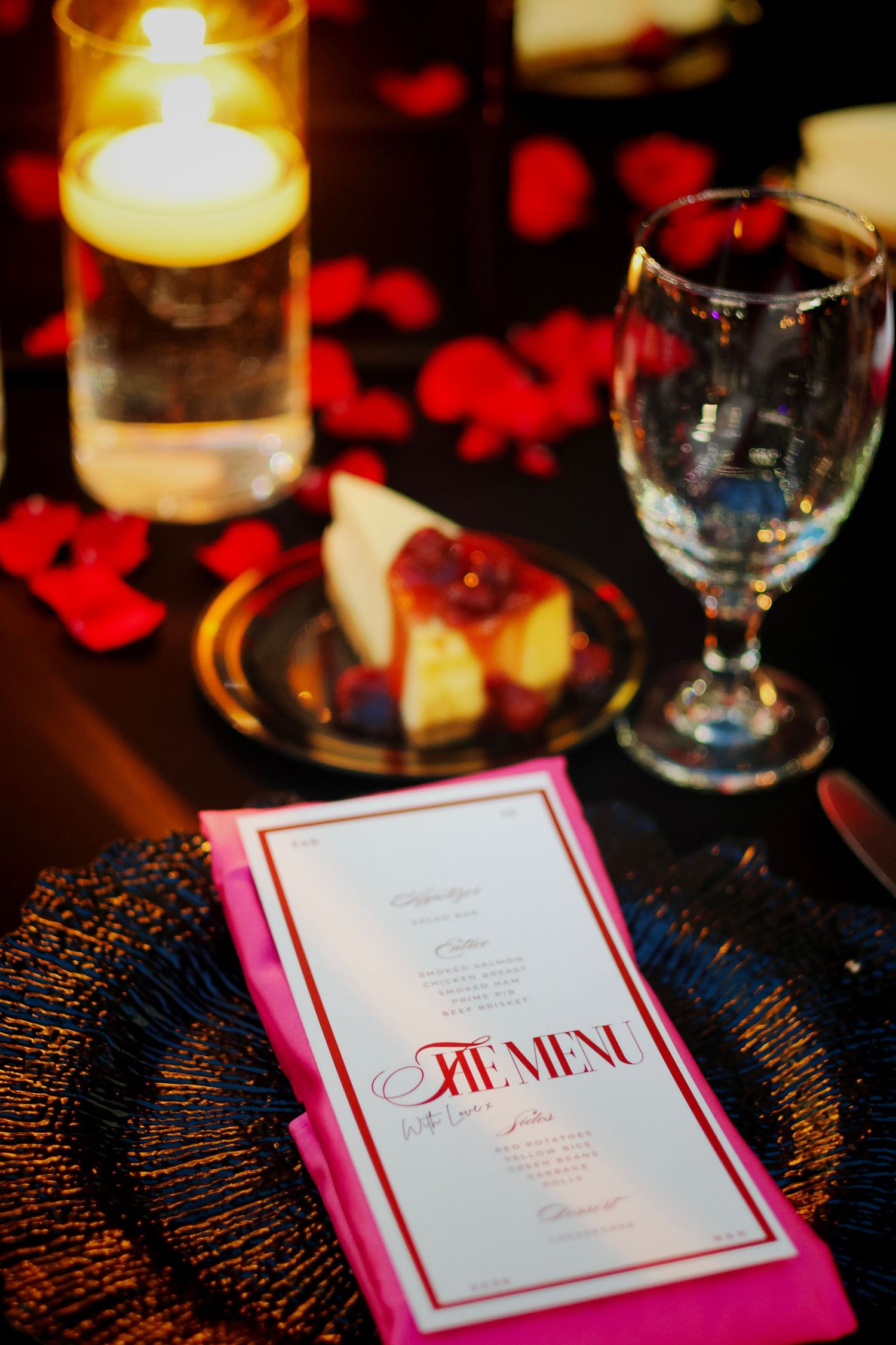 Elegant wedding table setting with pink menu card, lit candle, wine glass, and red rose petals for a romantic dinner.