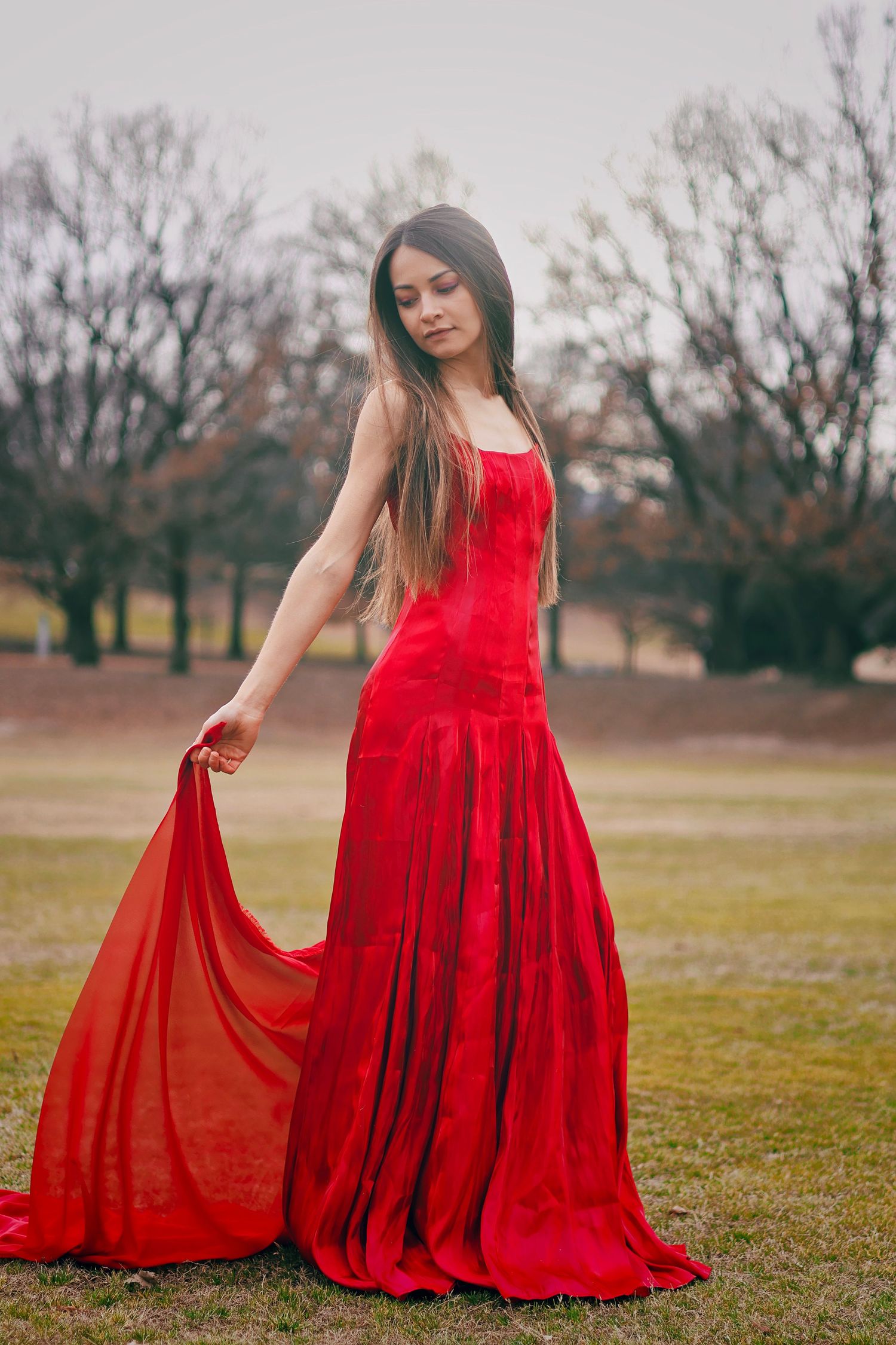 Woman in flowing red gown holds dress hem while standing gracefully in an outdoor park setting.