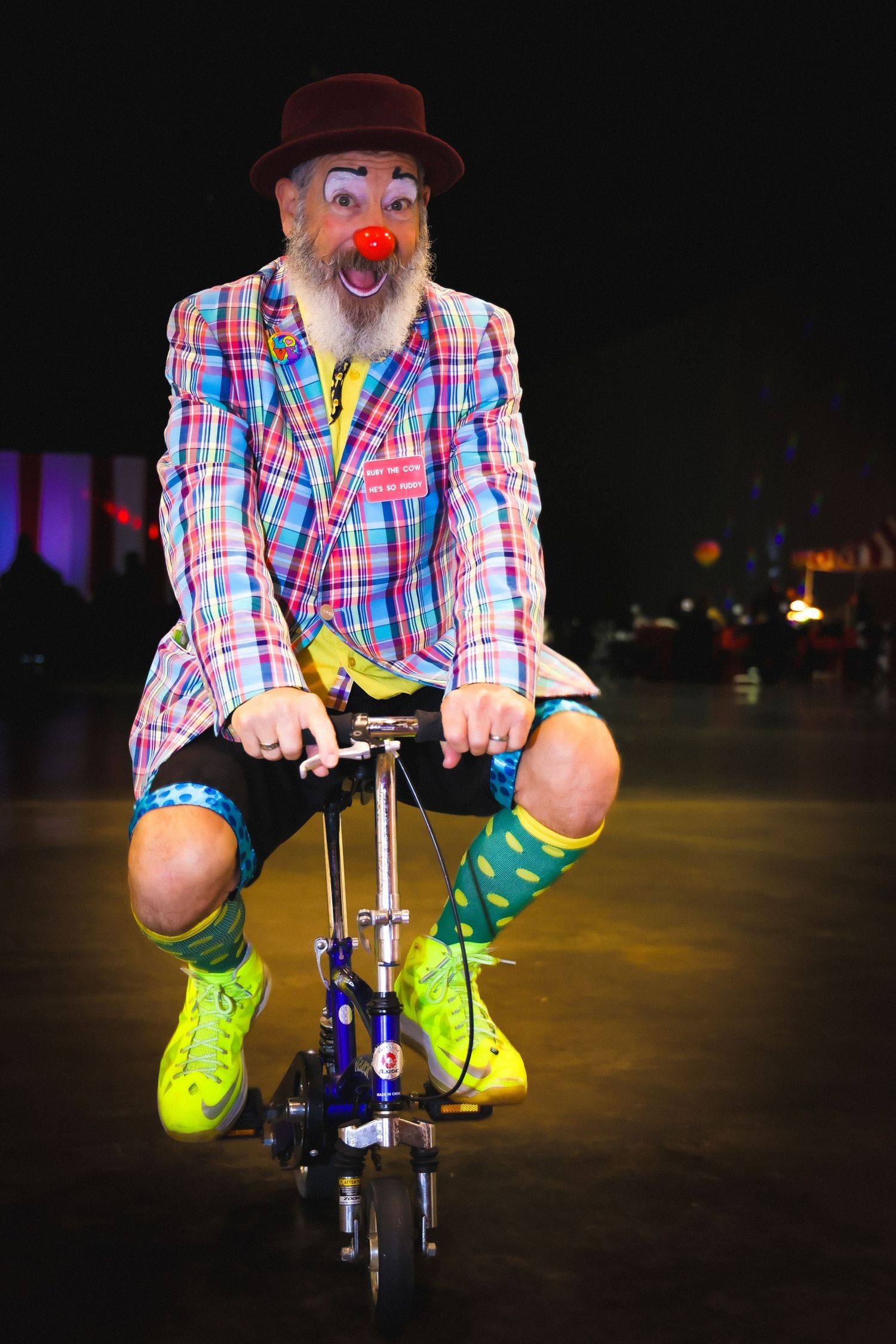 Elderly clown with red nose and beard rides tiny bicycle wearing colorful plaid jacket, hat, green socks, and yellow shoes.