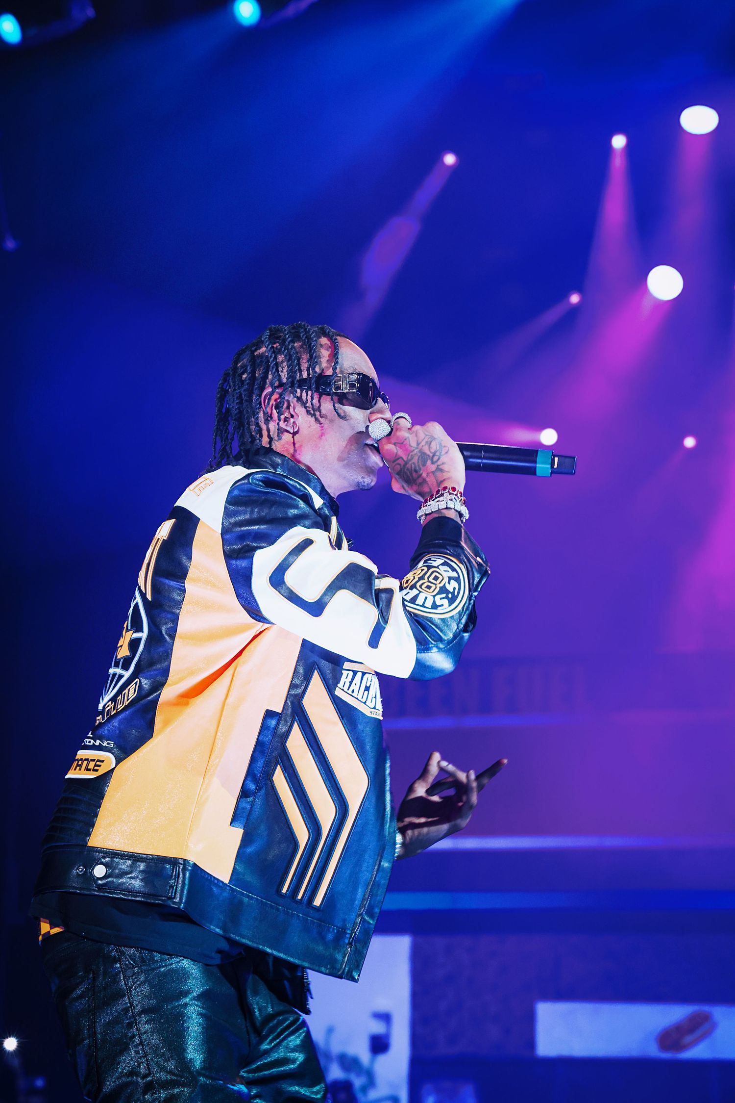 A rapper performing on stage with a microphone, wearing a black and yellow varsity jacket under blue stage lights.
