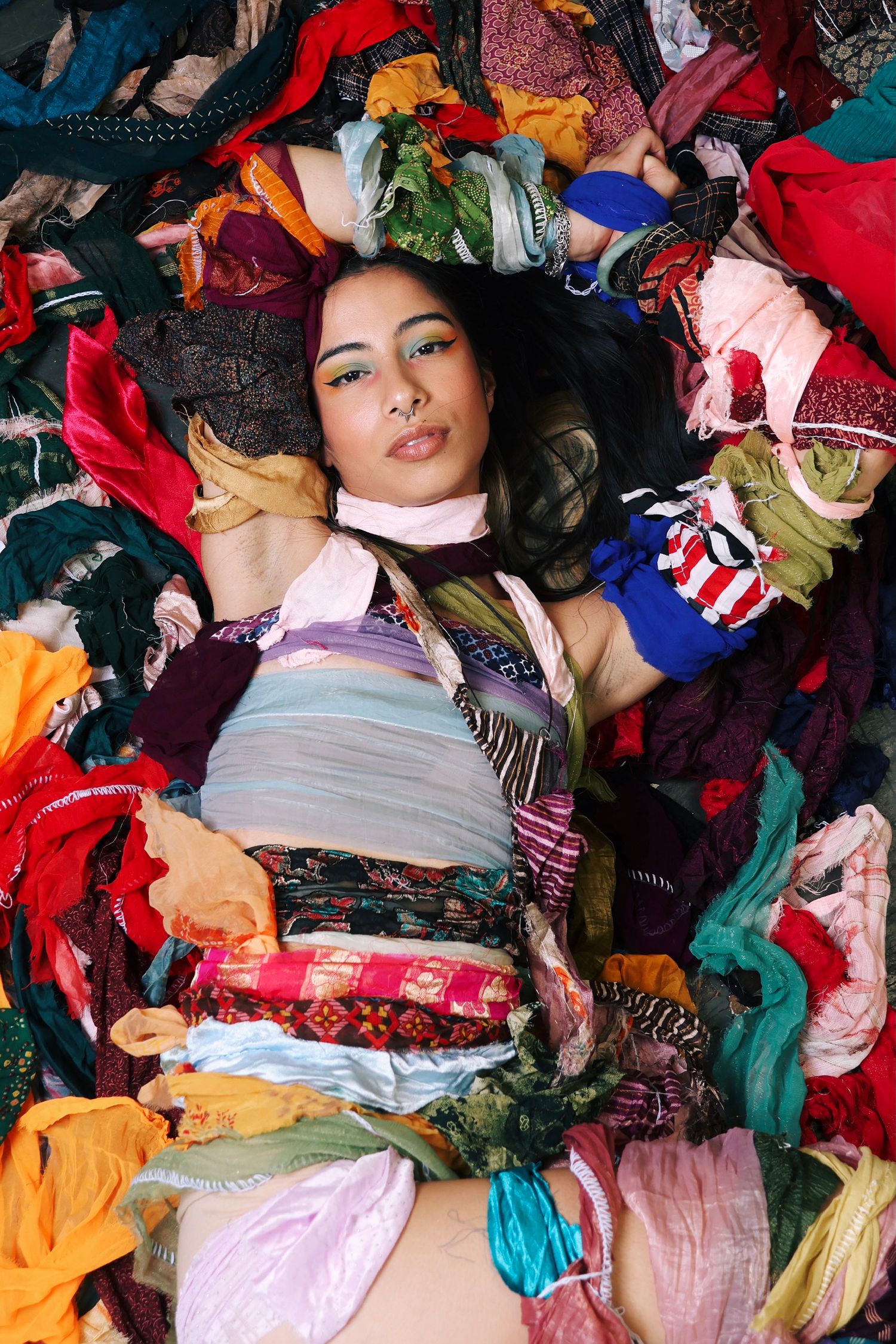 Young woman lying in a colorful pile of mixed fabric clothing scraps, wearing a patchwork outfit, fashion editorial style.