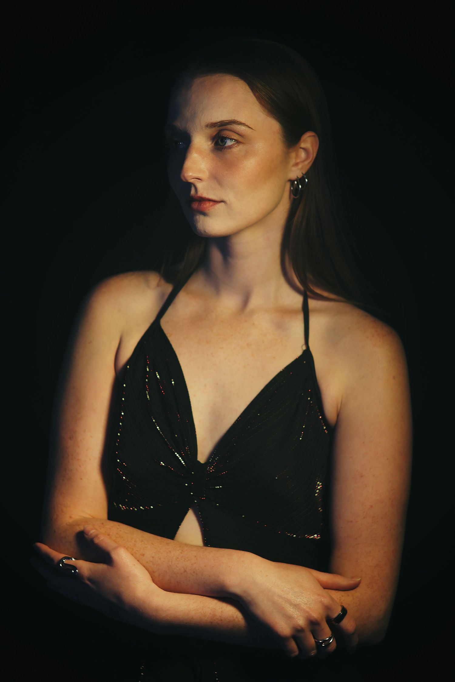 Young woman in black sequin halter top with arms crossed poses in dramatic low-key studio lighting.