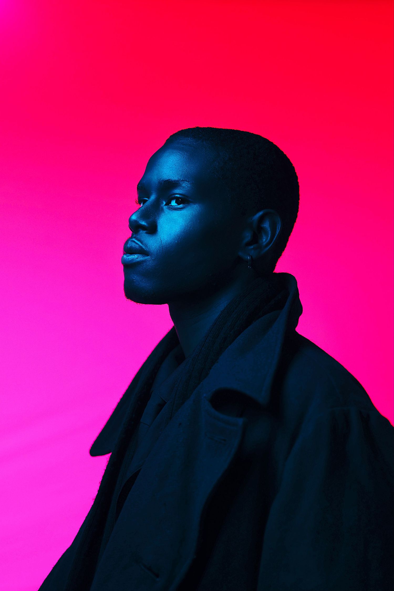 Profile portrait of Black woman in dark coat with blue and pink neon lighting contrast.