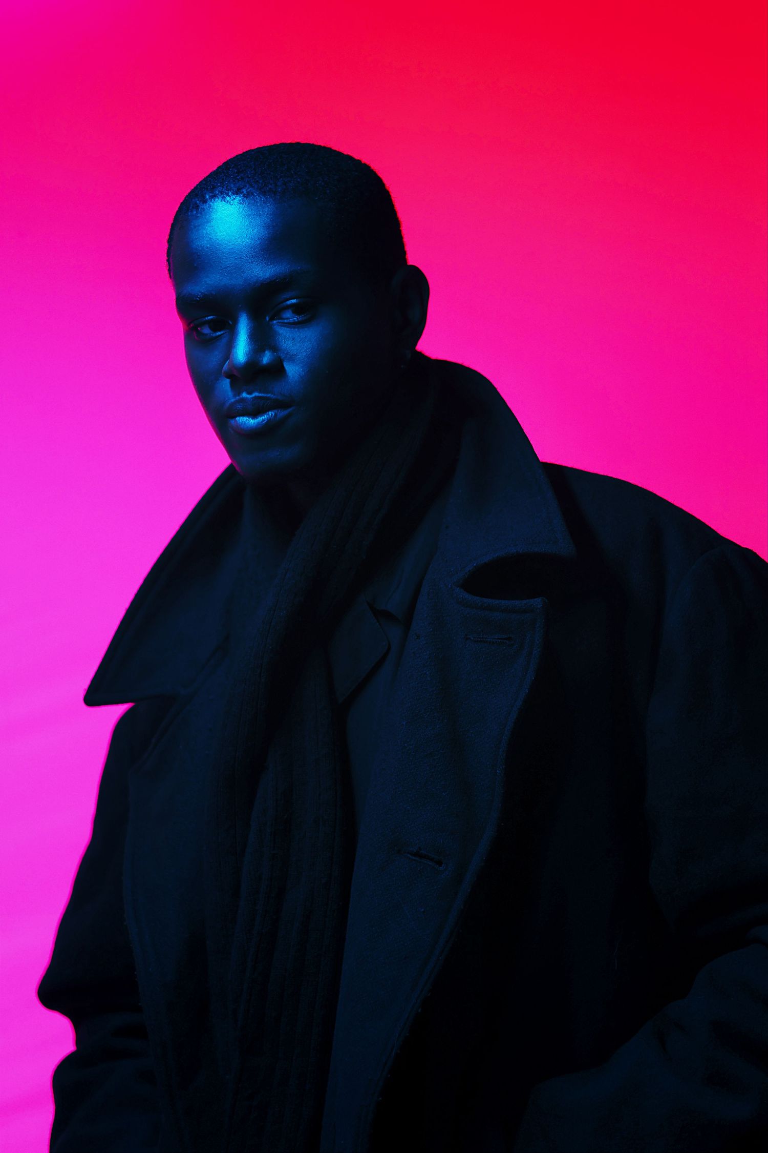 A Black man in a dark coat poses against a vibrant pink-magenta background with dramatic blue lighting.