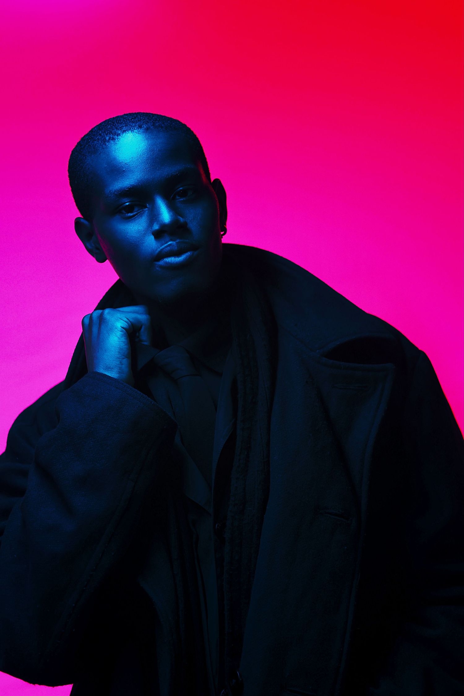 A young Black man in a black jacket poses against a vibrant pink-red background under dramatic blue neon lighting.