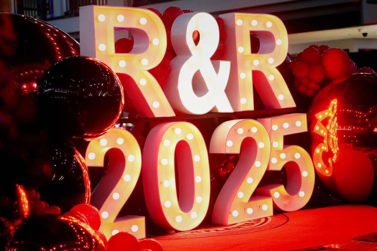 Illuminated marquee letters R&R 2025 with light bulbs, surrounded by red balloons and festive decorations.
