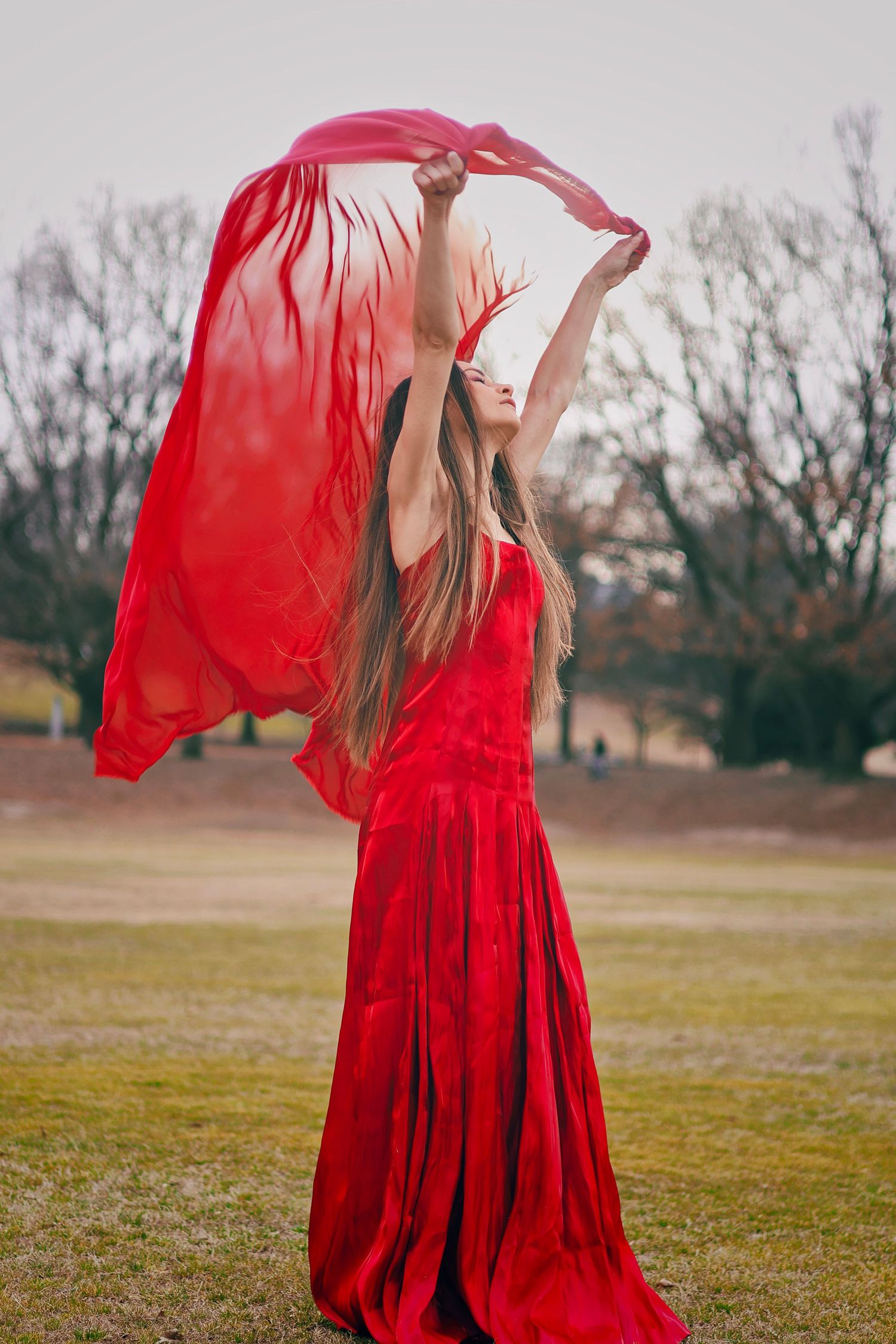 Woman in flowing red gown holds red fabric aloft in a park, her long hair cascading as the cloth billows in the wind.