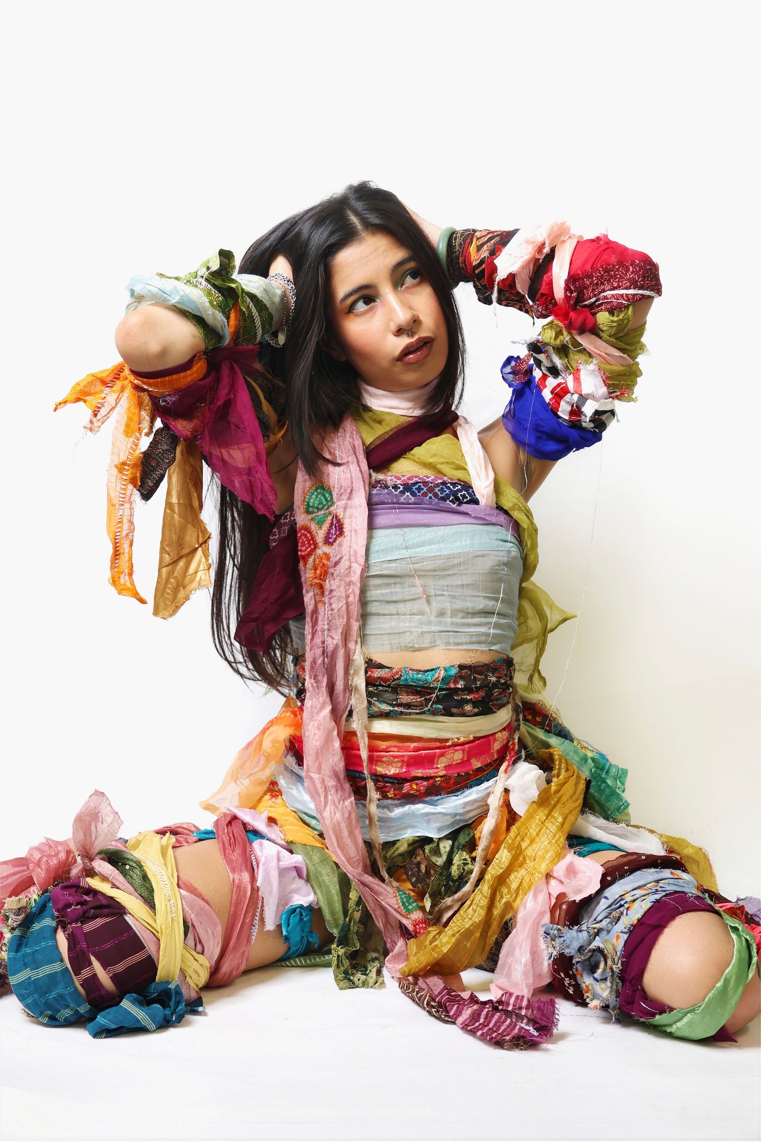 Young woman wrapped in colorful patchwork fabric strips sitting on white background in artistic fashion pose.