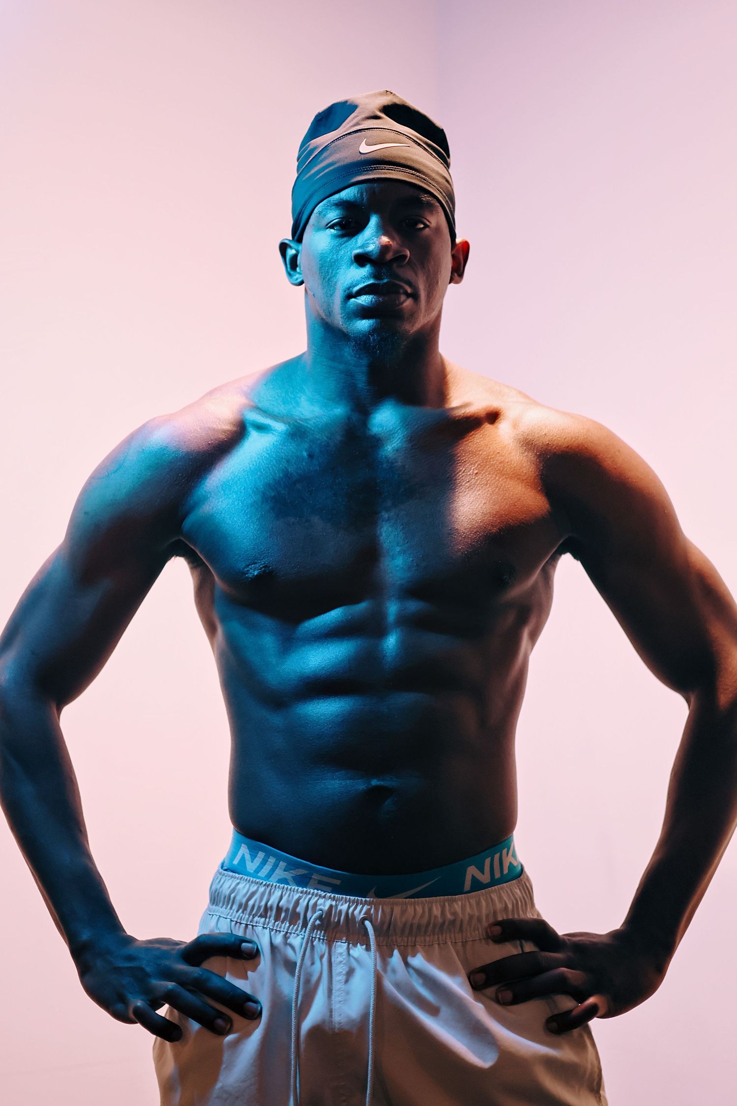 Muscular shirtless man wearing Nike headband and shorts posing confidently under blue studio lighting.