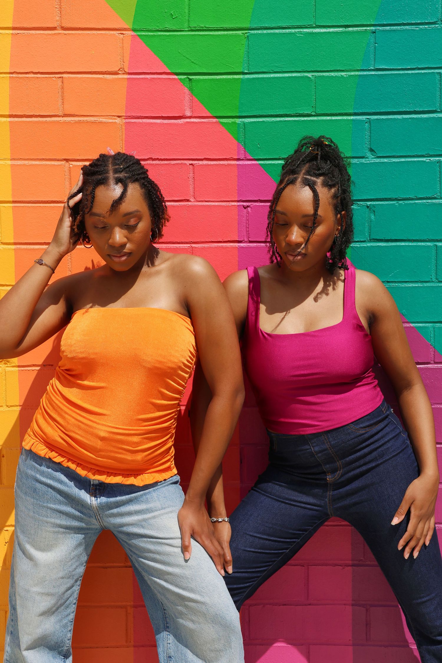 Two Black women in orange and pink tops posing against colorful rainbow brick wall mural.