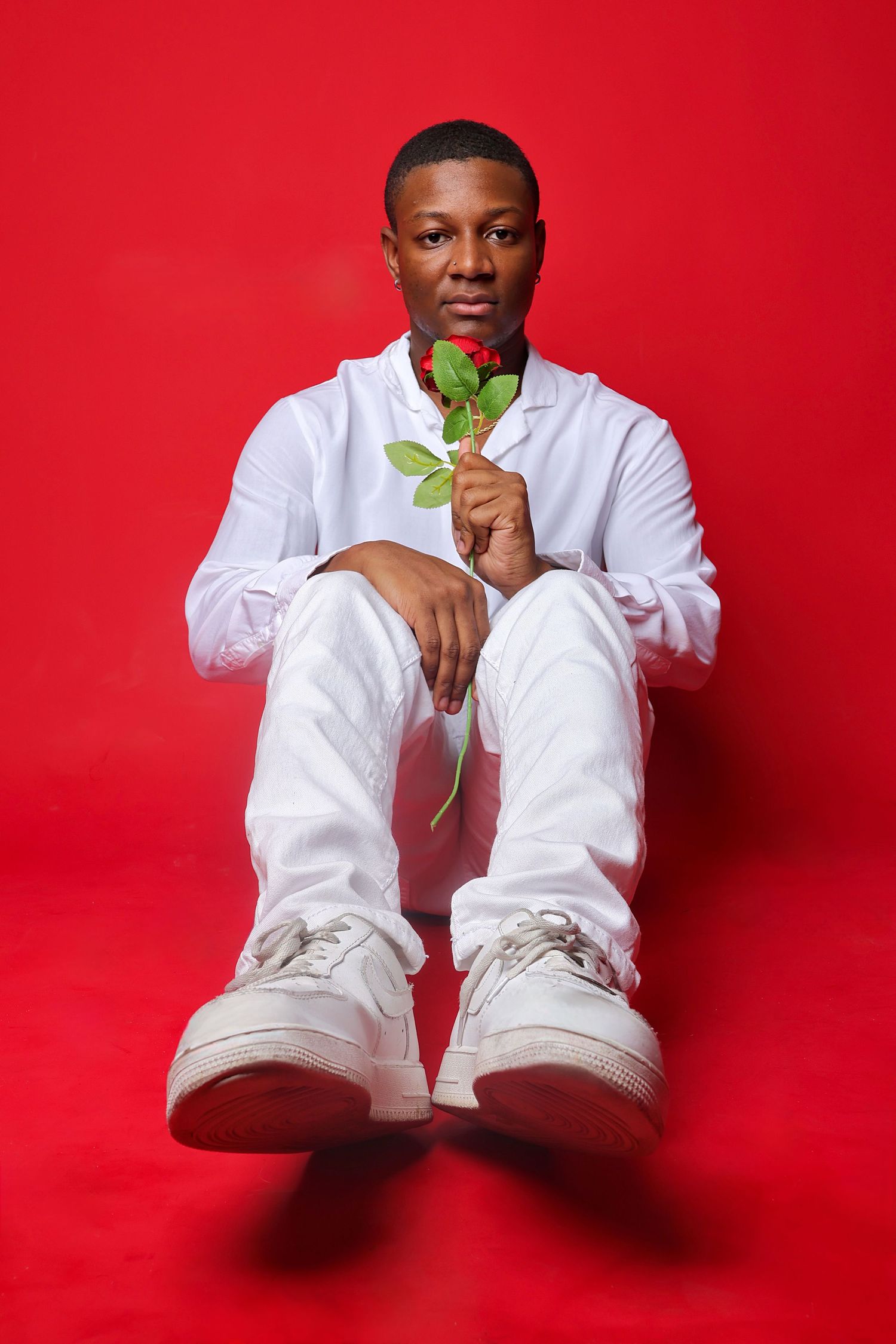 A young Black man in white outfit sits on red background holding a single red rose, looking directly at camera.