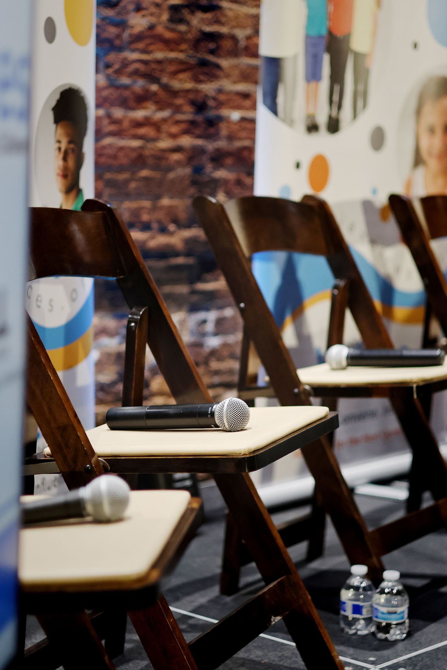 Wooden folding chairs with microphones on seats arranged in a row on a stage with colorful banners in the background.
