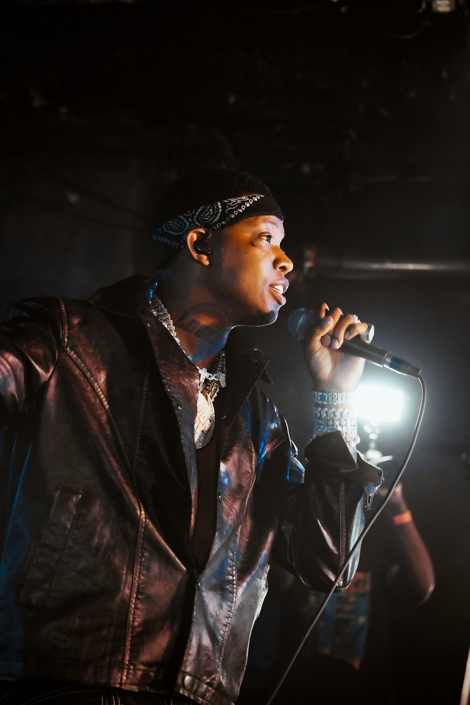 Young male rapper performing on stage, holding microphone, wearing leather jacket and bandana under dramatic spotlight.