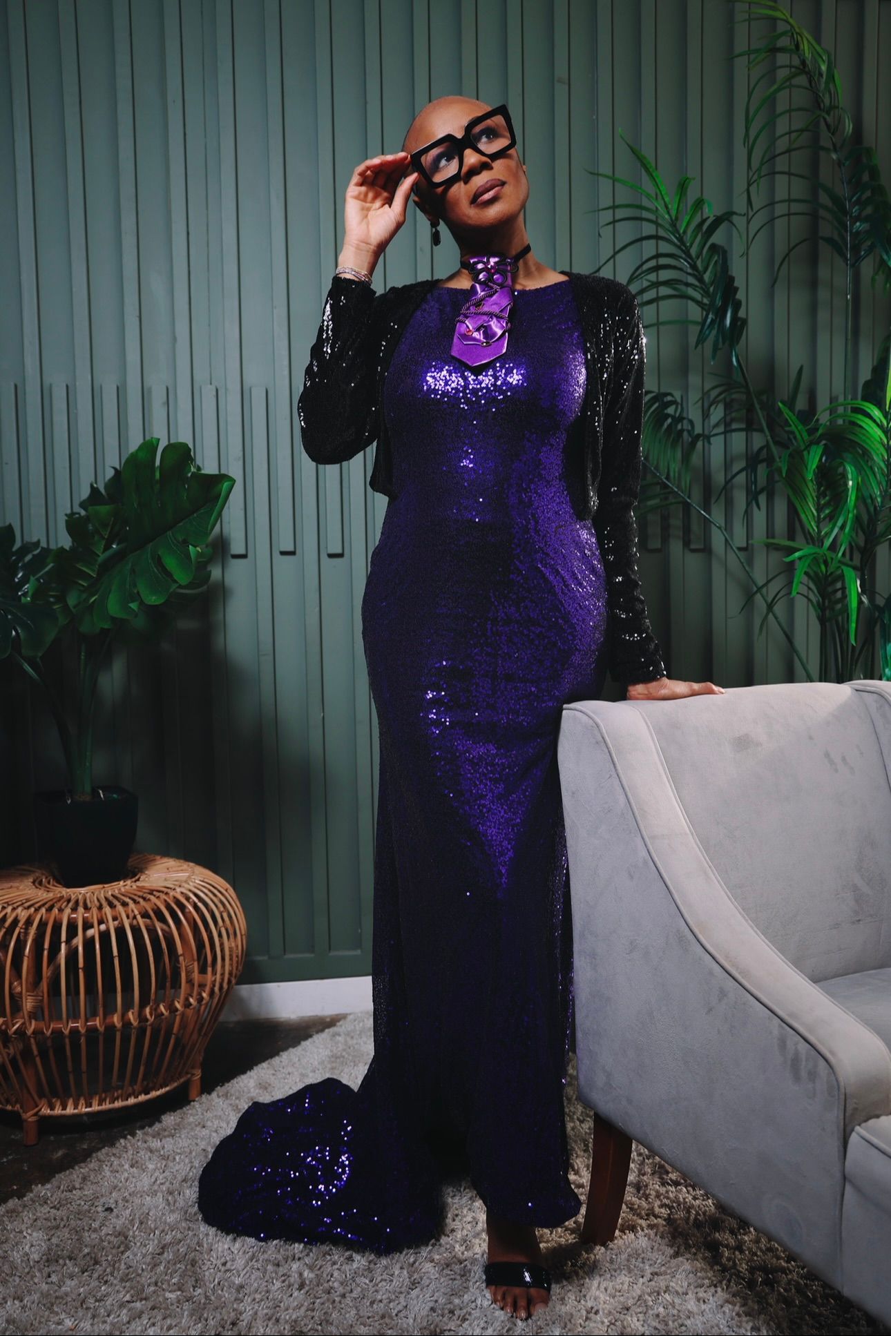 A bald Black woman poses in a purple sequin gown and black glasses beside a gray sofa in a green-walled room.
