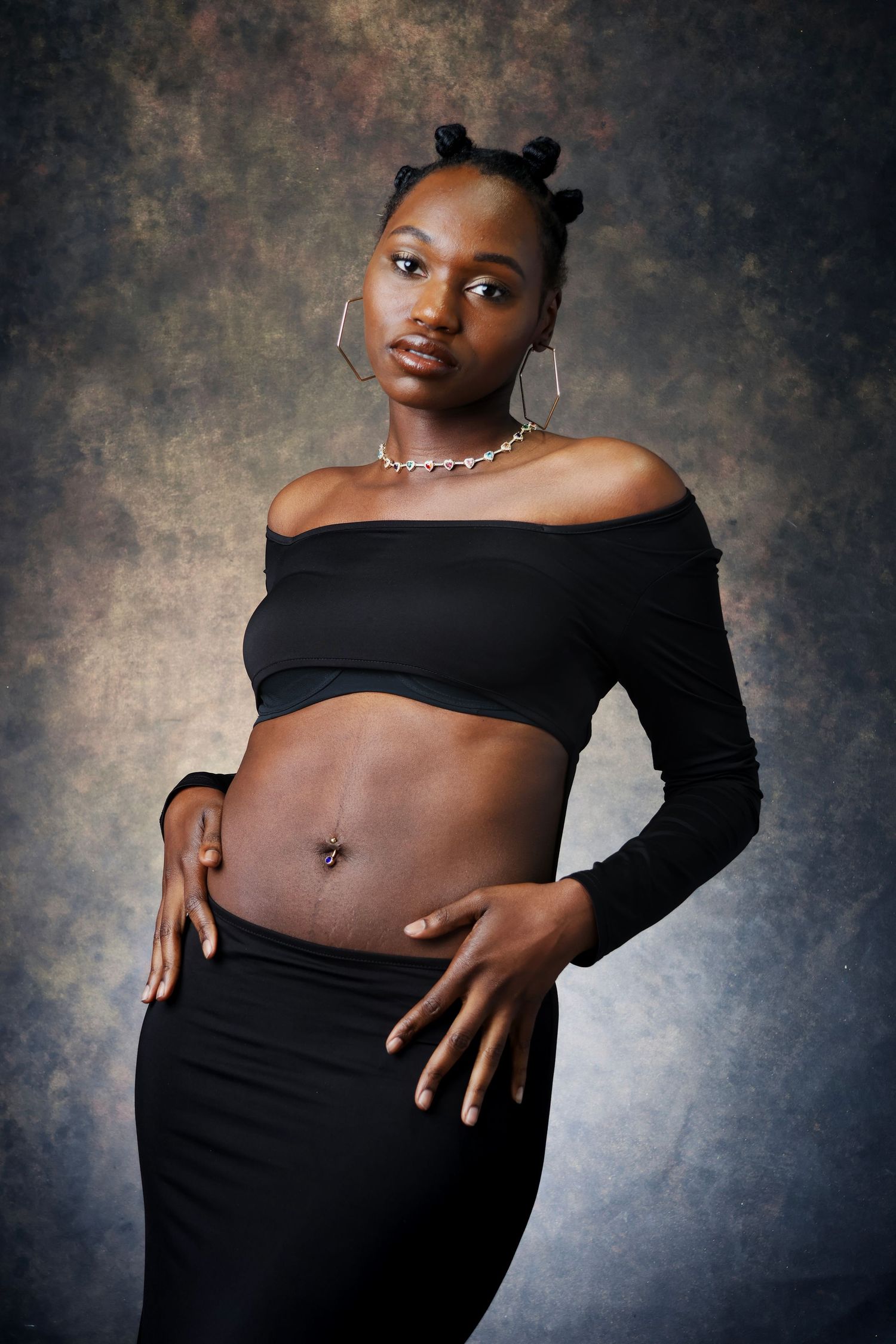 Young Black woman in black off-shoulder crop top and skirt posing against textured studio backdrop.