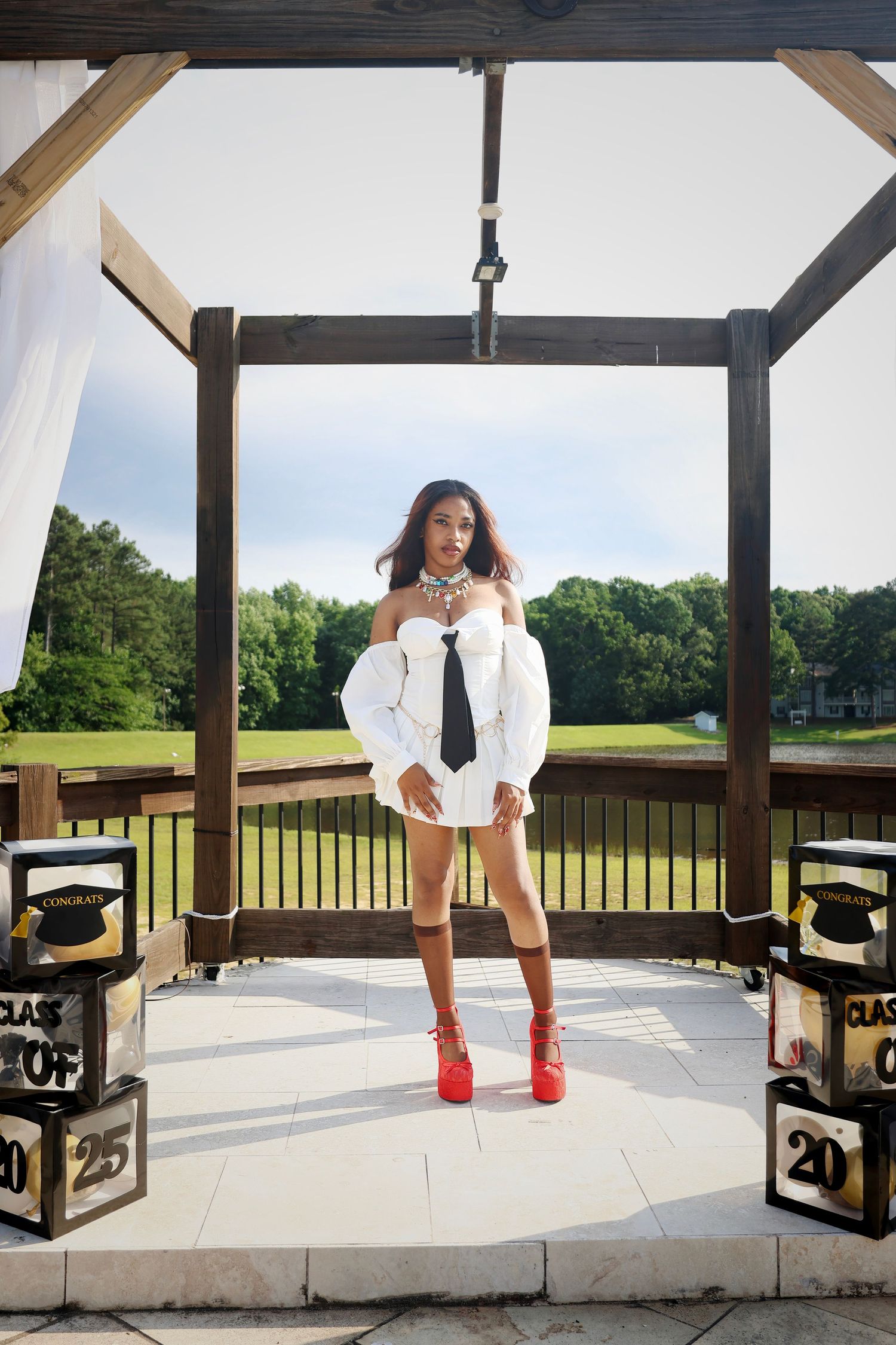 Young woman in white off-shoulder shirt with black tie and red heels poses in gazebo with Class of 2025 graduation decor.