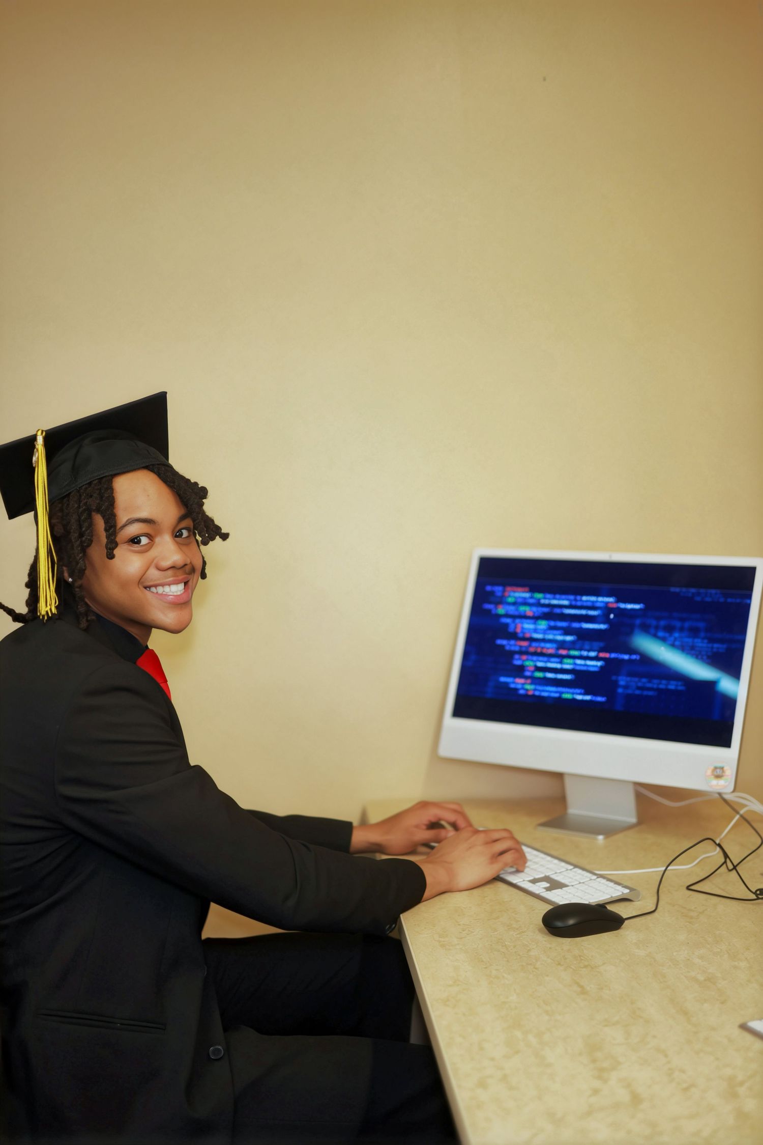 Graduate in cap and gown smiling at desk with computer displaying code, celebrating academic achievement.